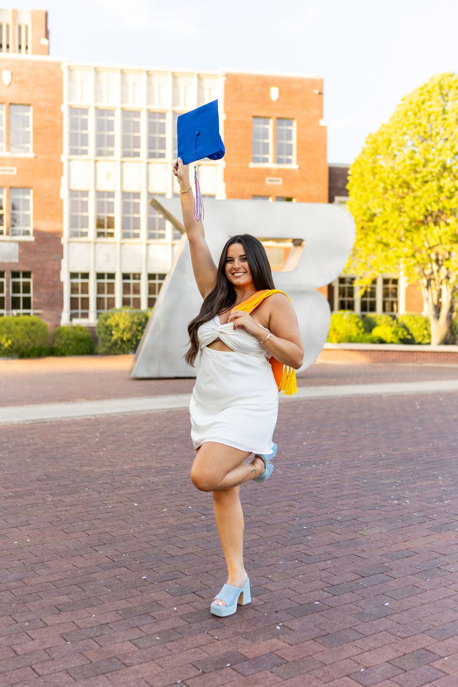 Graduate in white dress jumping and celebrating on brick courtyard with school building in background.