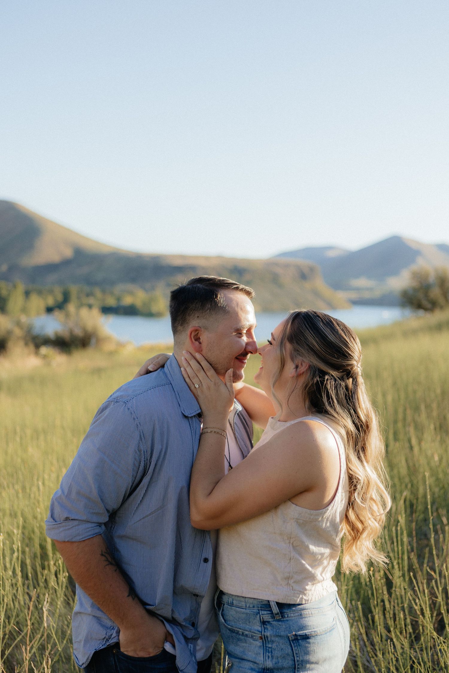 Couple sharing an intimate moment in tall grass with mountains and lake in the background.