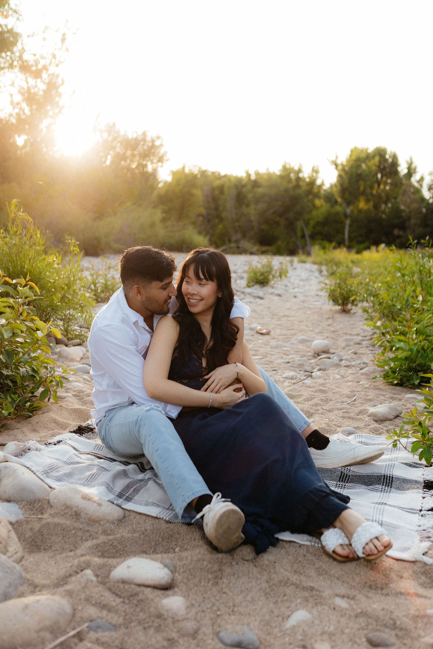 A couple sits intimately on a blanket at the beach during golden hour sunset, surrounded by wild flowers and sand.