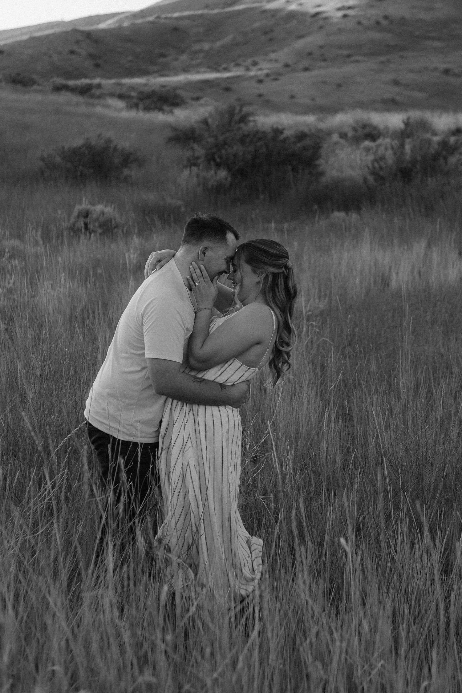 Romantic black and white photo of a couple embracing in tall grass with mountains in background.