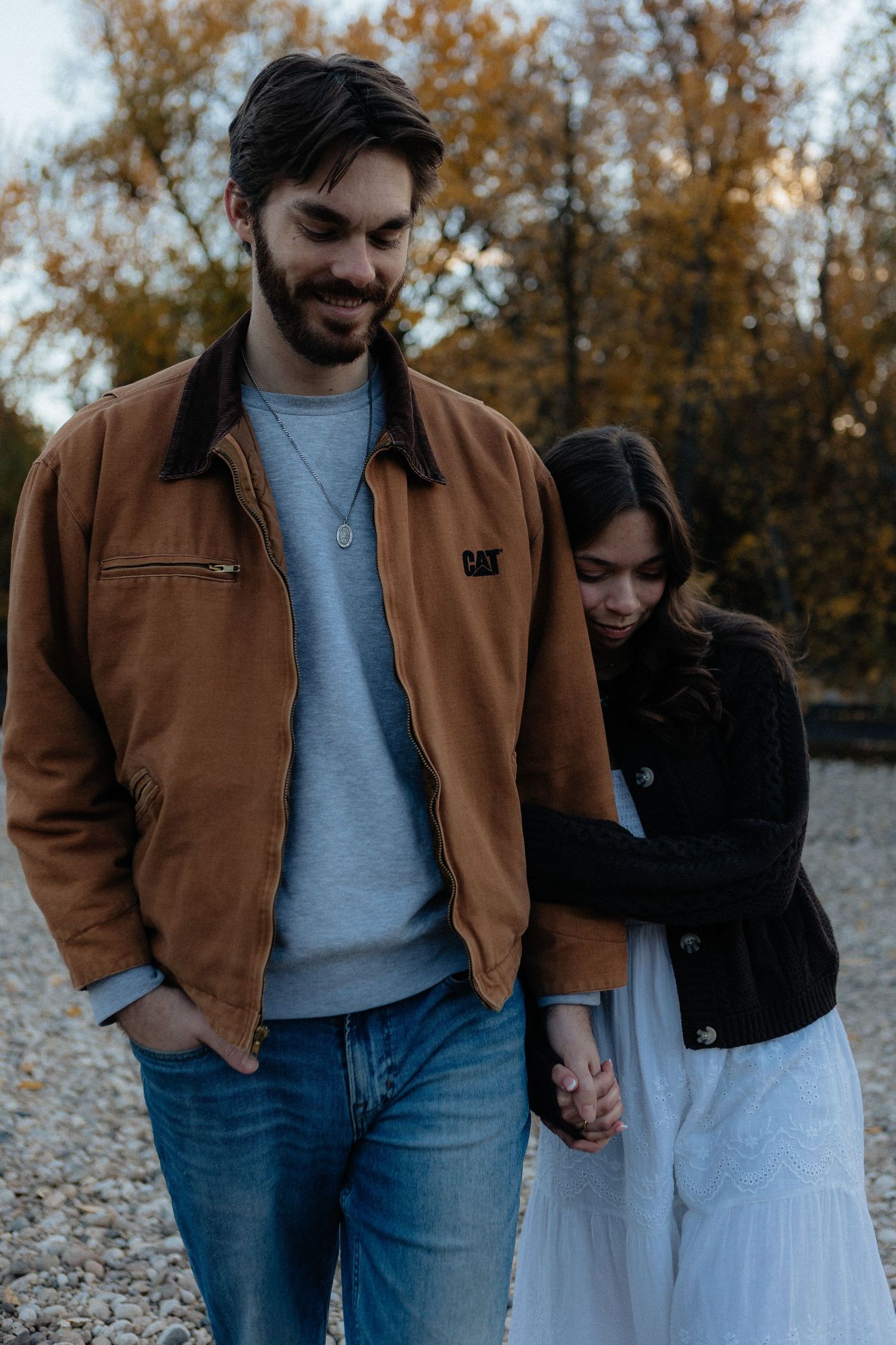 A couple in casual attire walks together on an autumn day, with the person wearing a brown jacket and jeans.