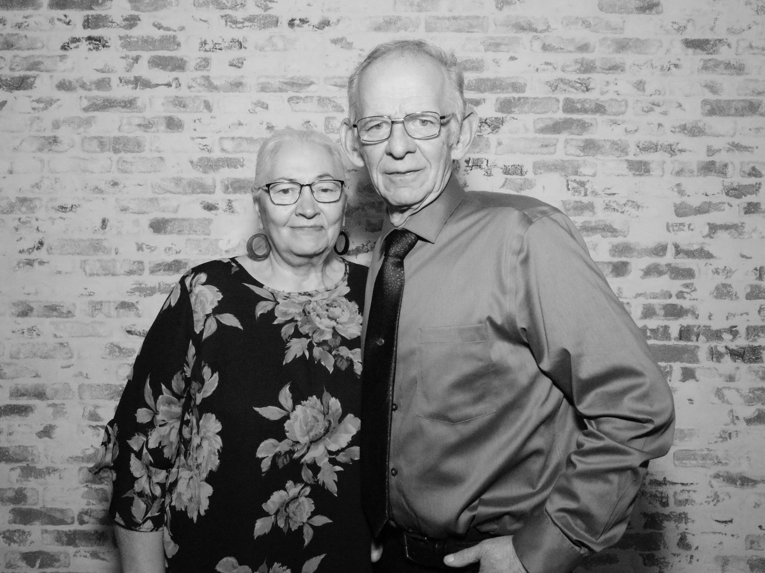Couple in formal attire smiling together against rustic brick wall backdrop.