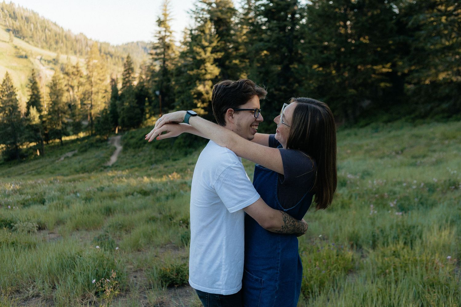 A couple embraces in a scenic mountain meadow during golden hour with evergreen trees in the background.