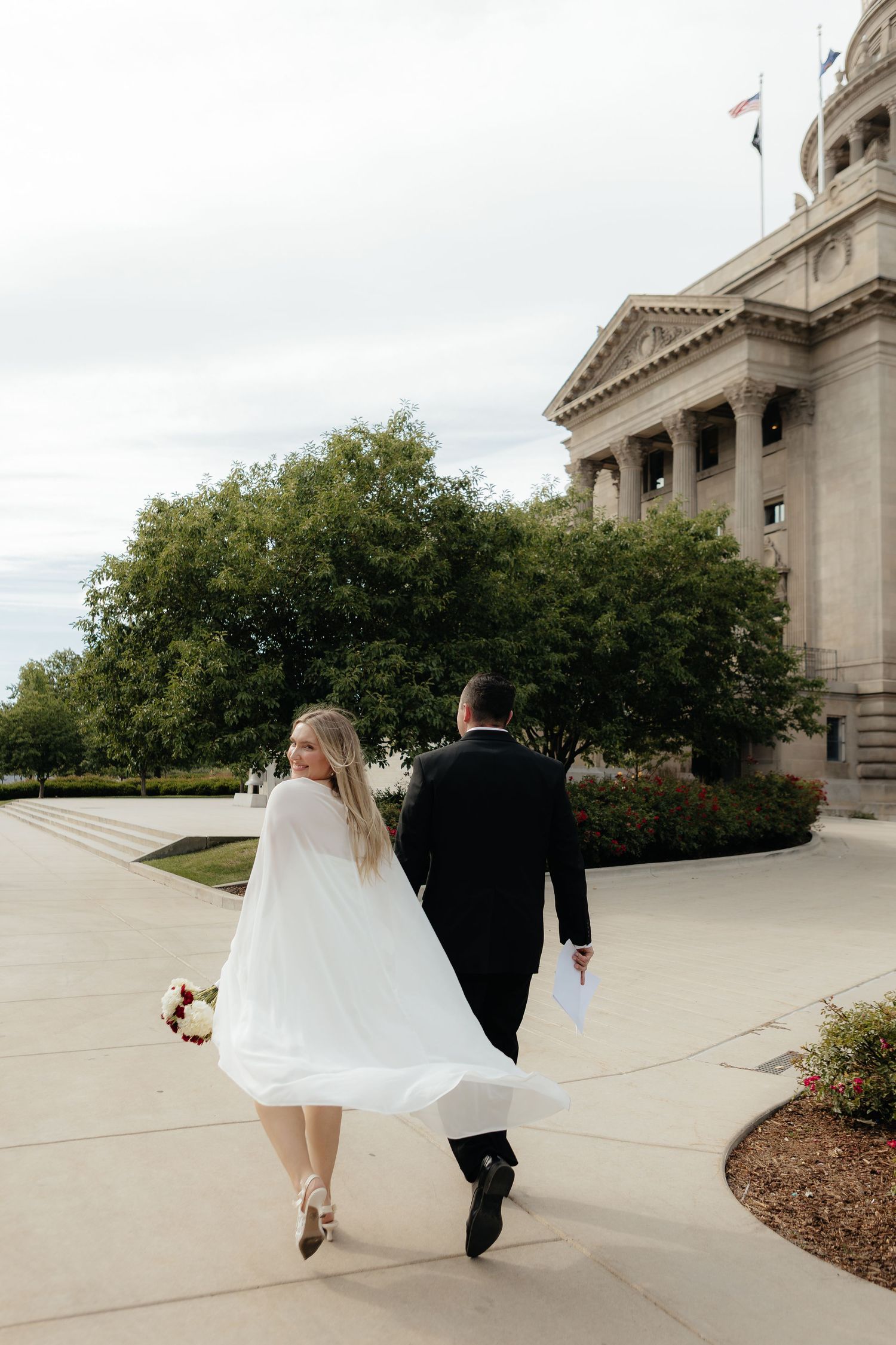 A couple in formal wedding attire walks along a curved concrete path outside a grand building with columns.