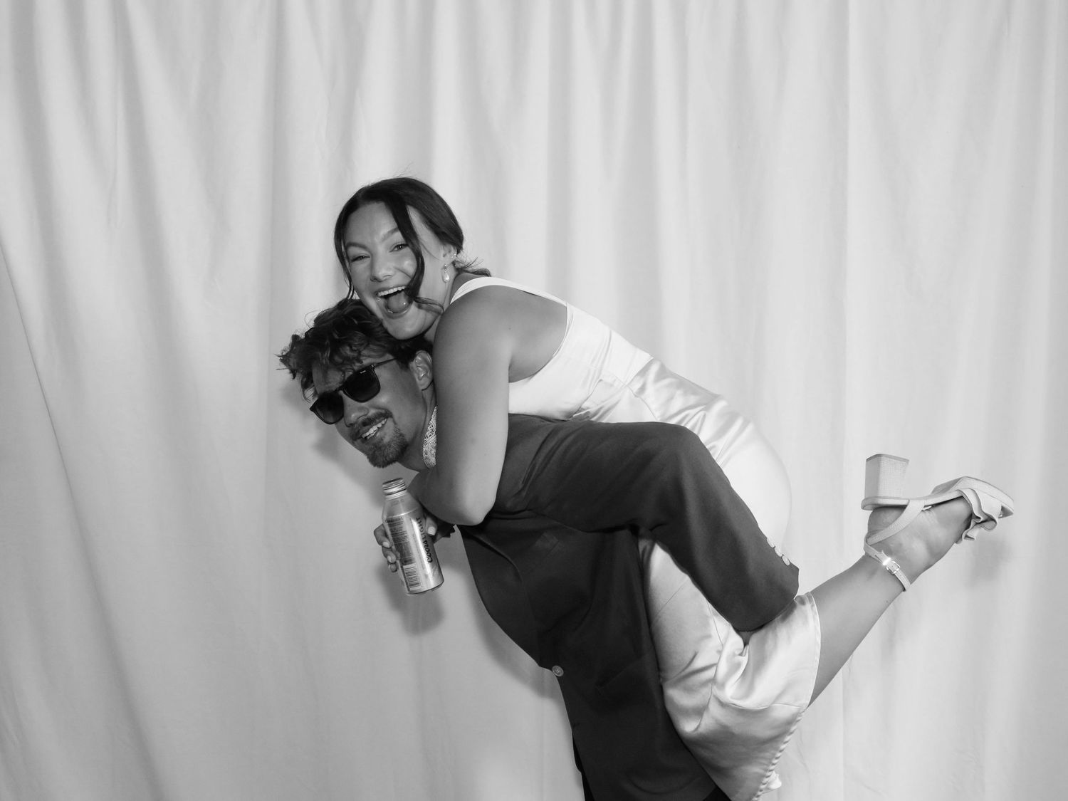 Black and white photo booth picture of a couple laughing, man giving woman a piggyback ride at a formal event.