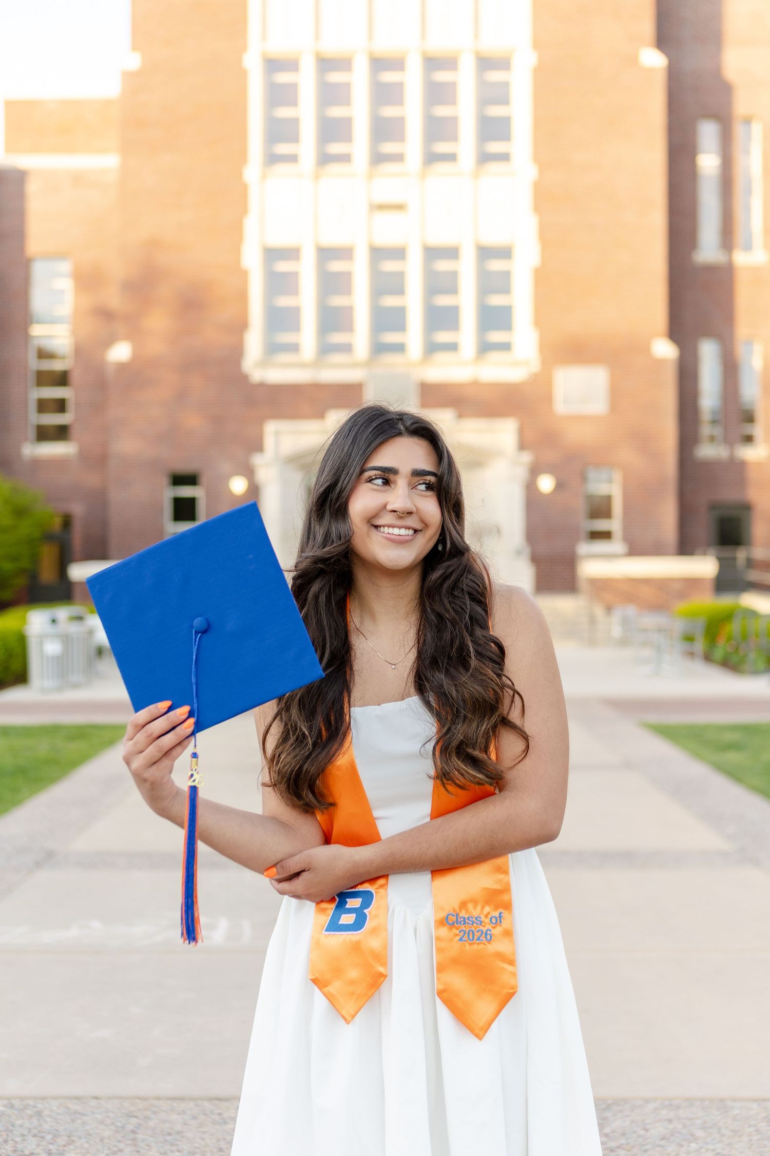 Smiling female graduate holds blue cap wearing white dress and orange sash in front of brick university building.