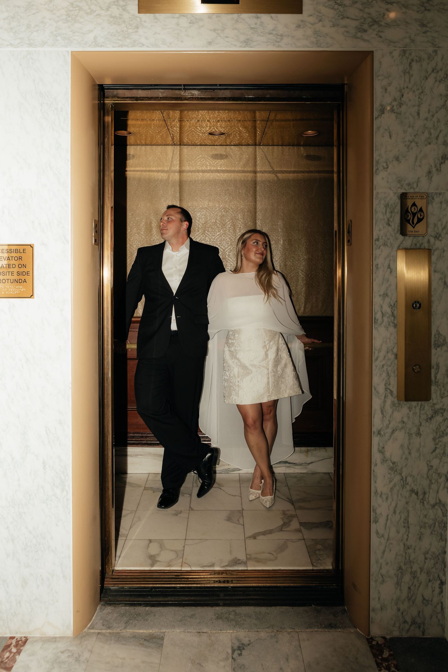 Two people in wedding attire pose together inside an elegant marble elevator with mirrored walls.