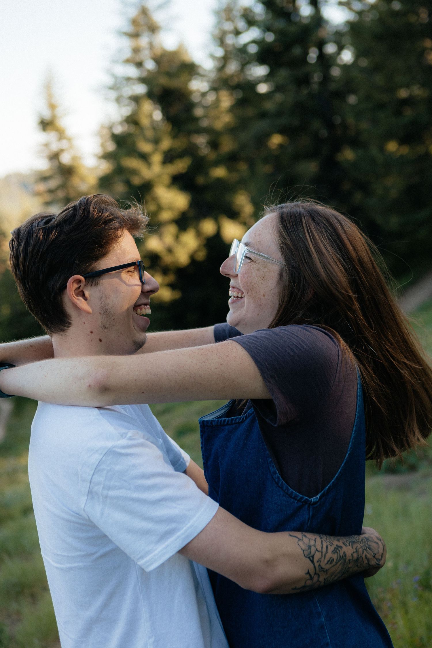 A couple embraces and playfully spins around together in a sunny outdoor field with evergreen trees in the background.