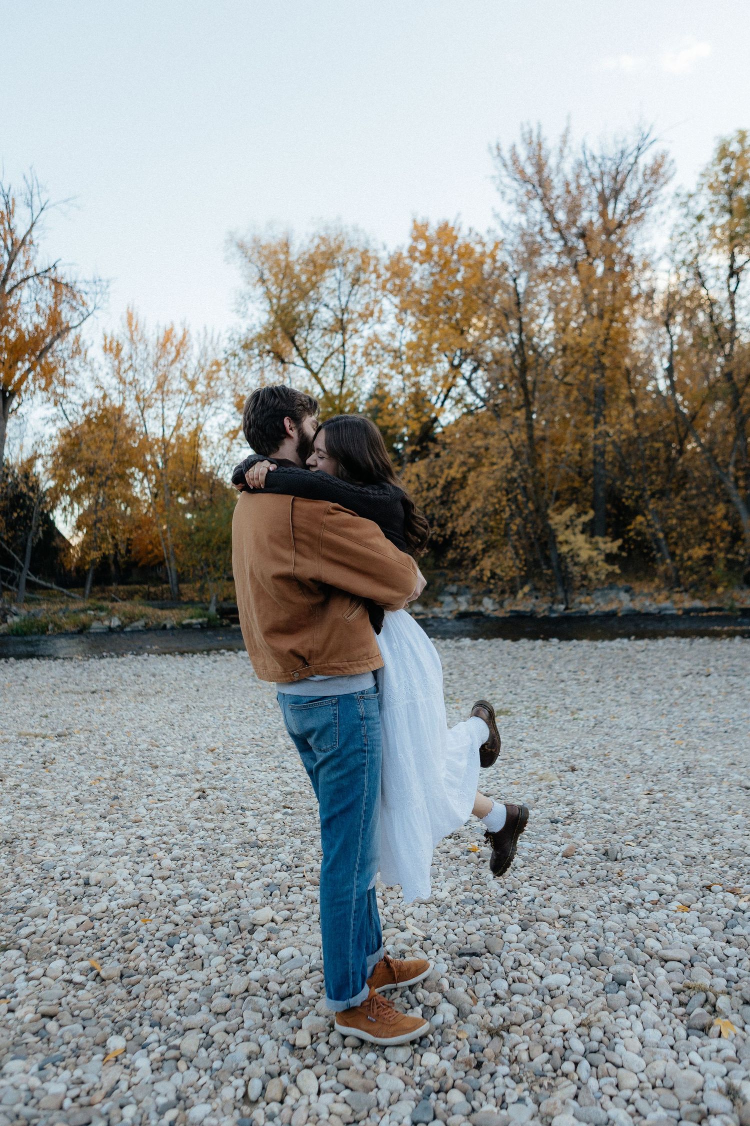Playful couple spins and embraces on rocky beach with autumn trees in background.