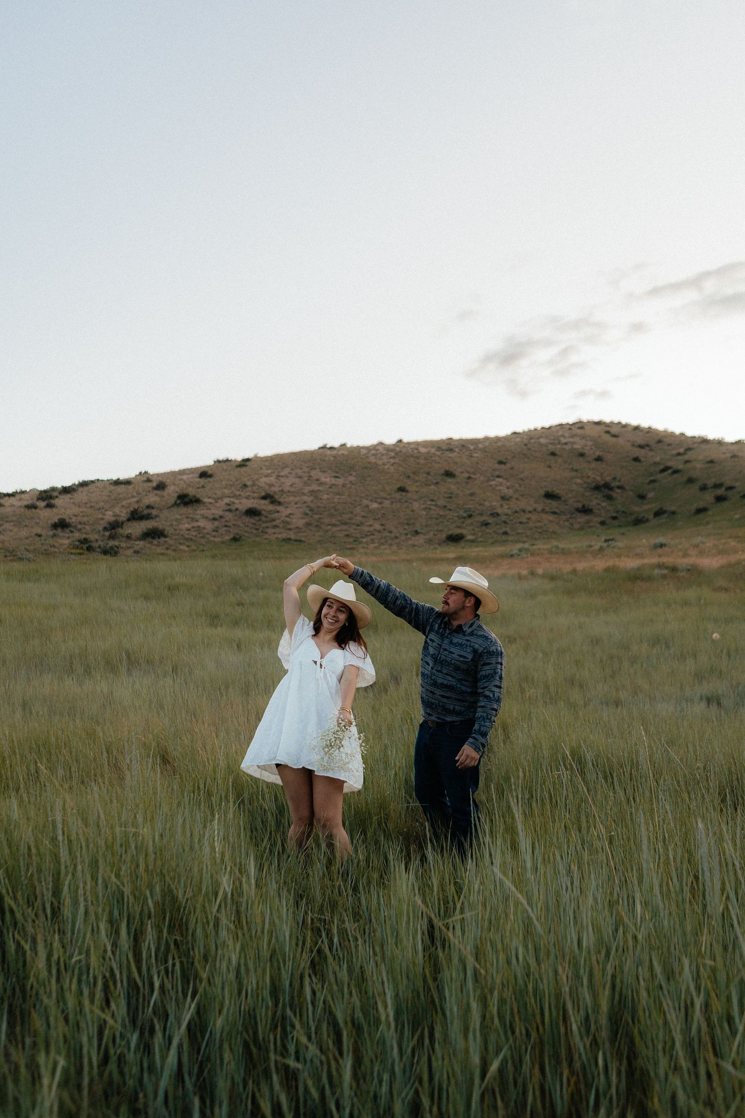 A couple dances together in a field of tall grass with rolling hills in the background at dusk.
