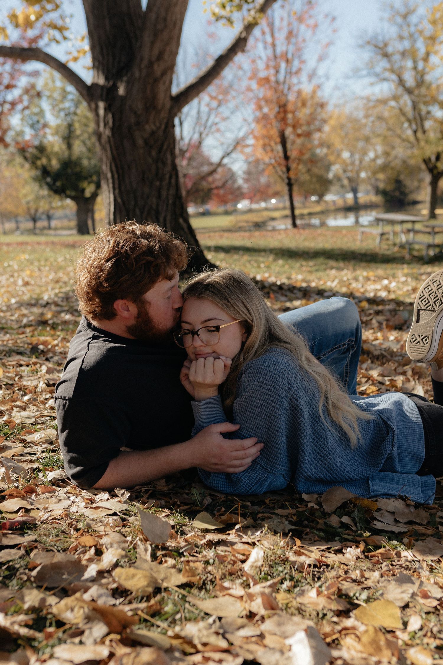 Couple lying together on autumn leaves under a large tree in a park.
