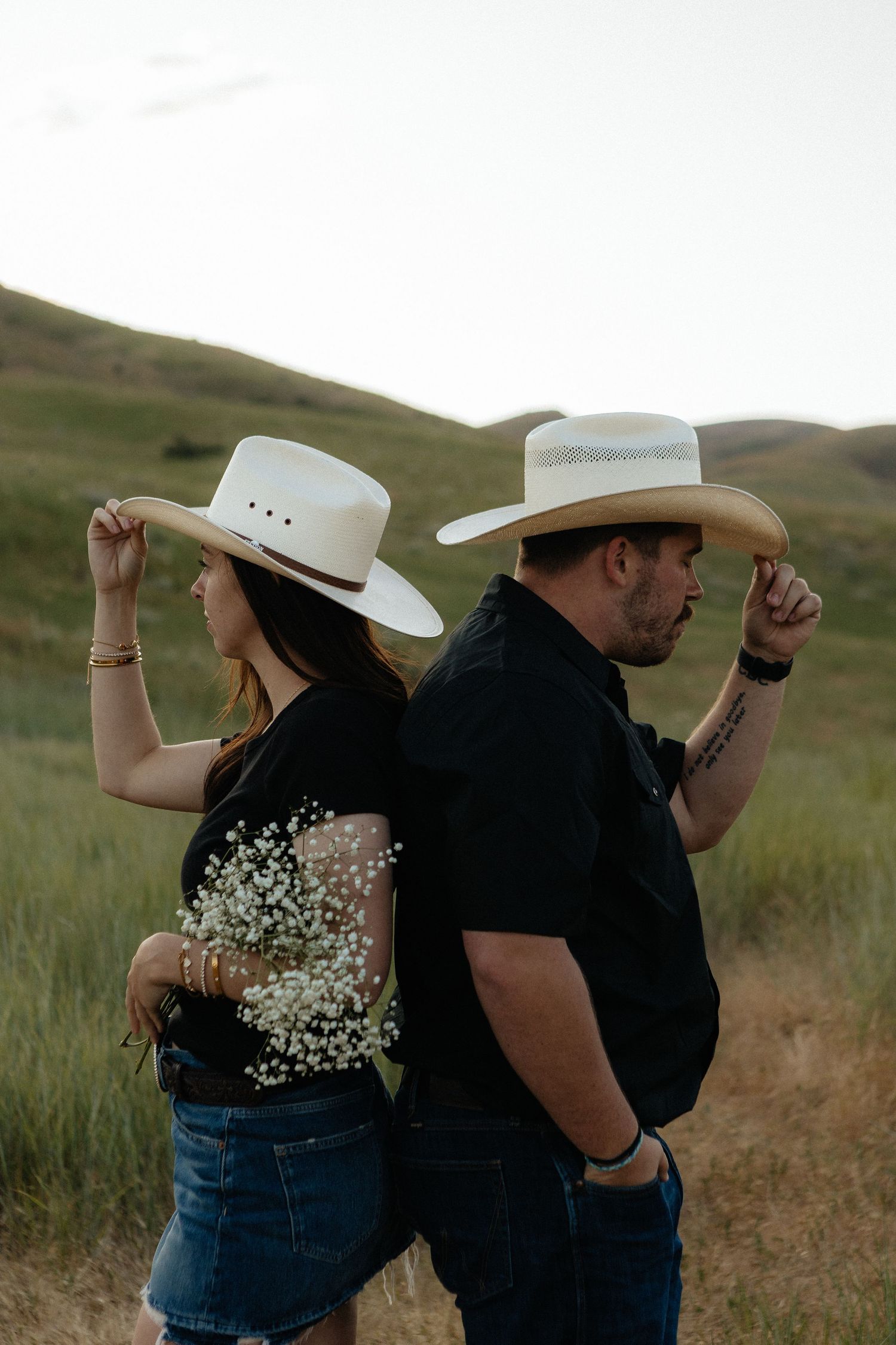 A couple in matching white cowboy hats and blue jeans dance together in a grassy field at sunset.