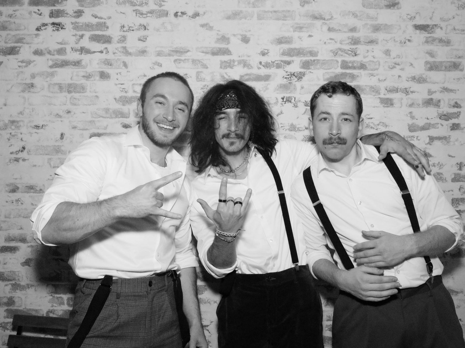 Group wearing suspenders and formal attire posing playfully against brick wall.