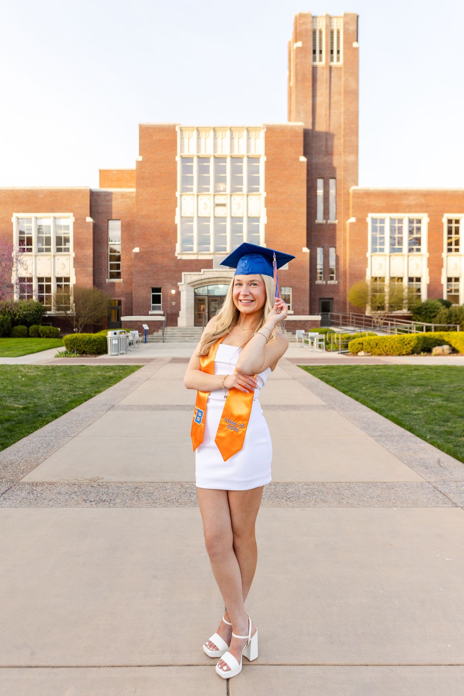 Smiling female graduate in white dress, blue cap, and orange sash poses in front of a brick university building.