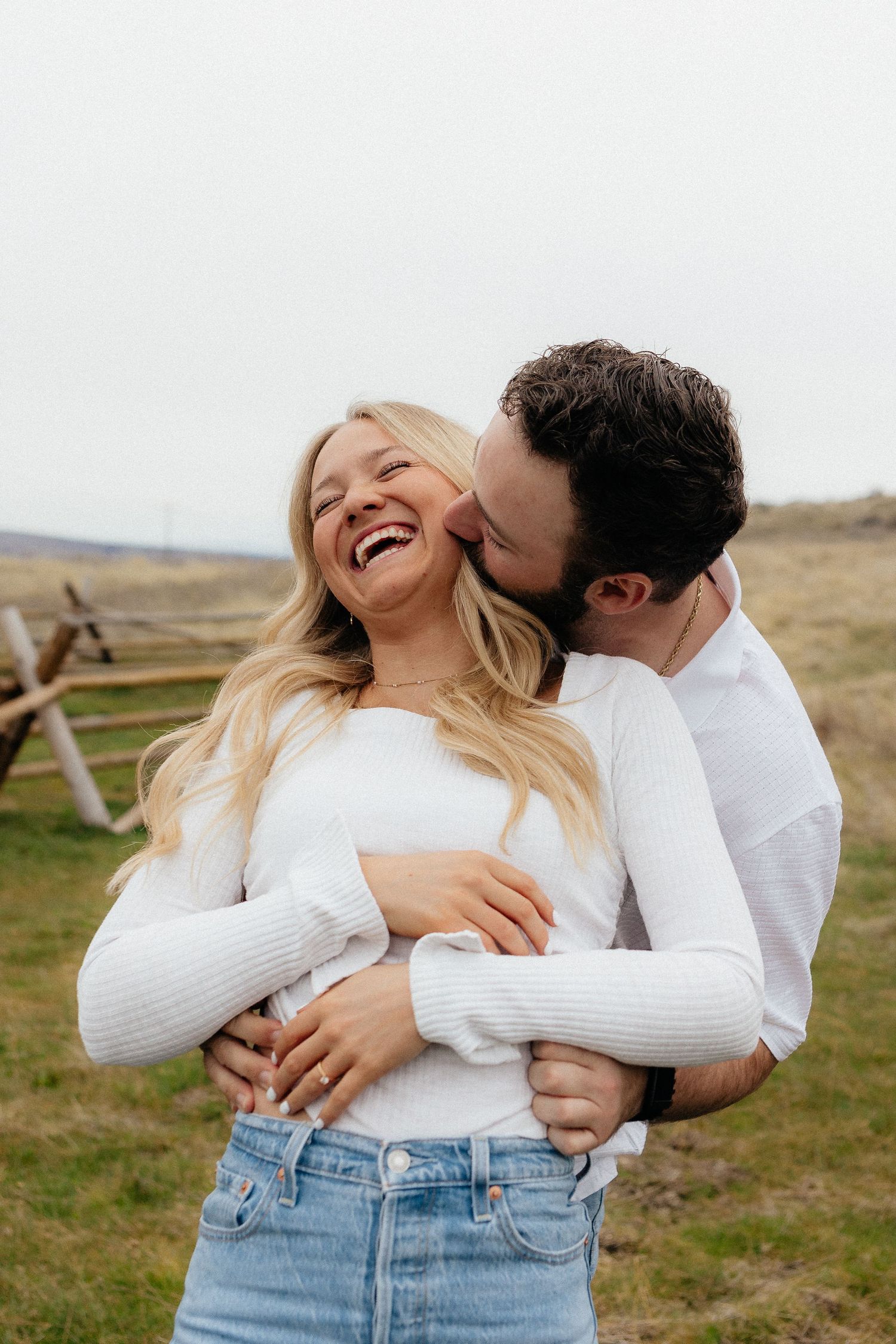 A couple in white tops embraces outdoors, the man kissing the woman's cheek as she laughs joyfully in a field.