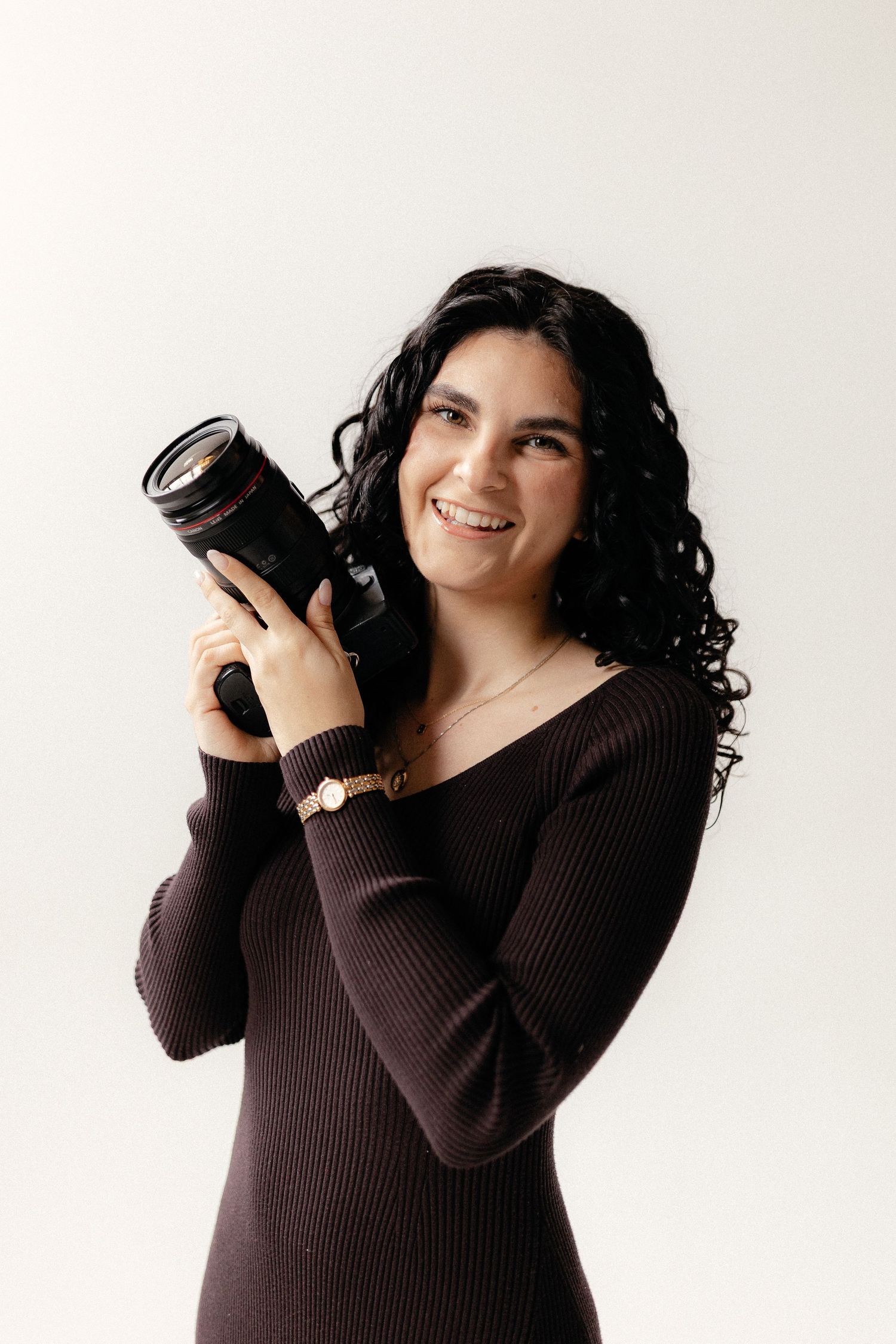A photographer in a brown ribbed sweater poses playfully with a camera against a white background.