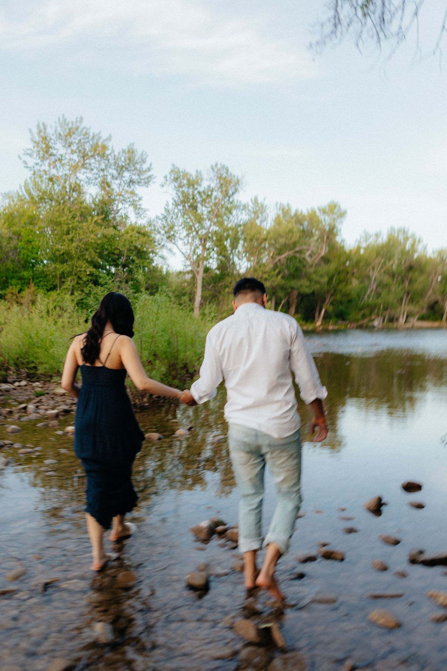 A couple holding hands while walking barefoot through shallow water with rocks along a peaceful lake shore.