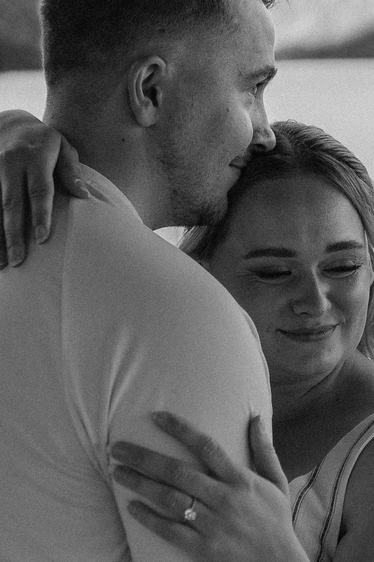 Intimate black and white portrait of a couple embracing with a sparkly engagement ring visible.