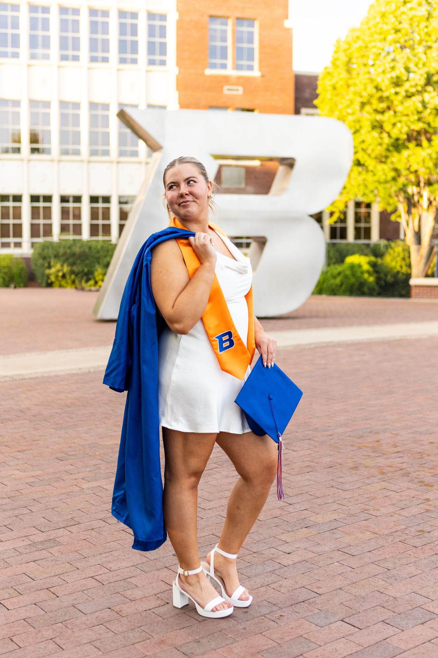 Graduate in white dress and blue regalia poses with diploma on campus brick courtyard.