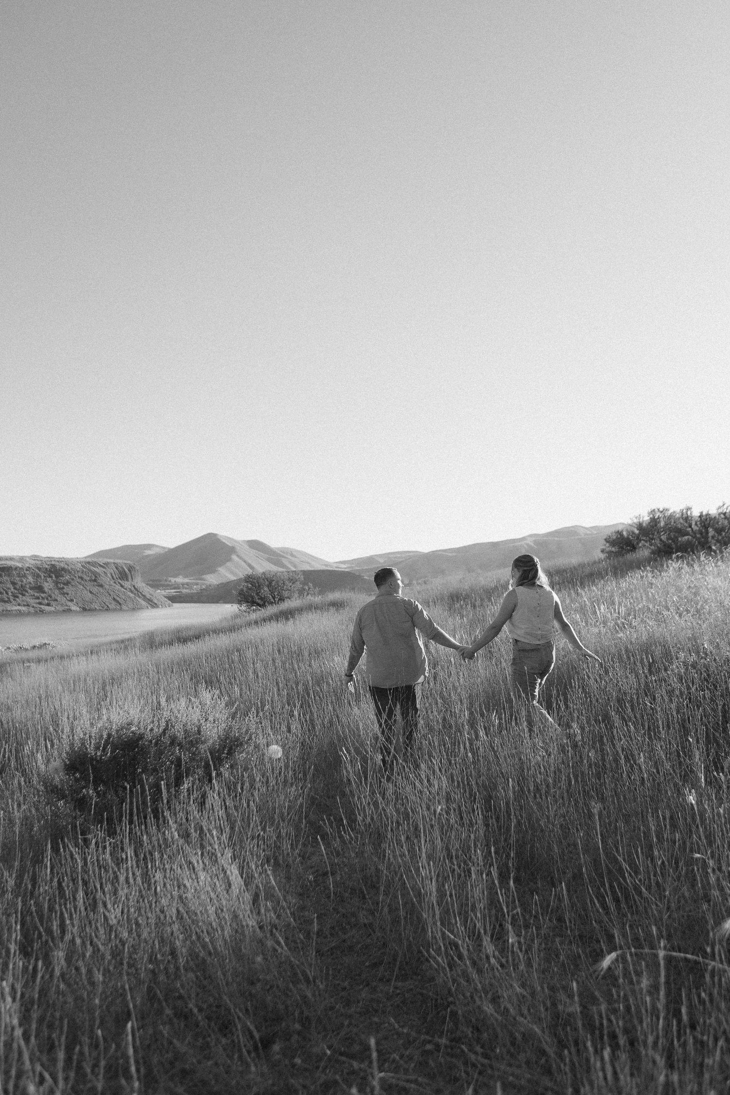 Two people hold hands while walking through tall grass with mountains in the background.