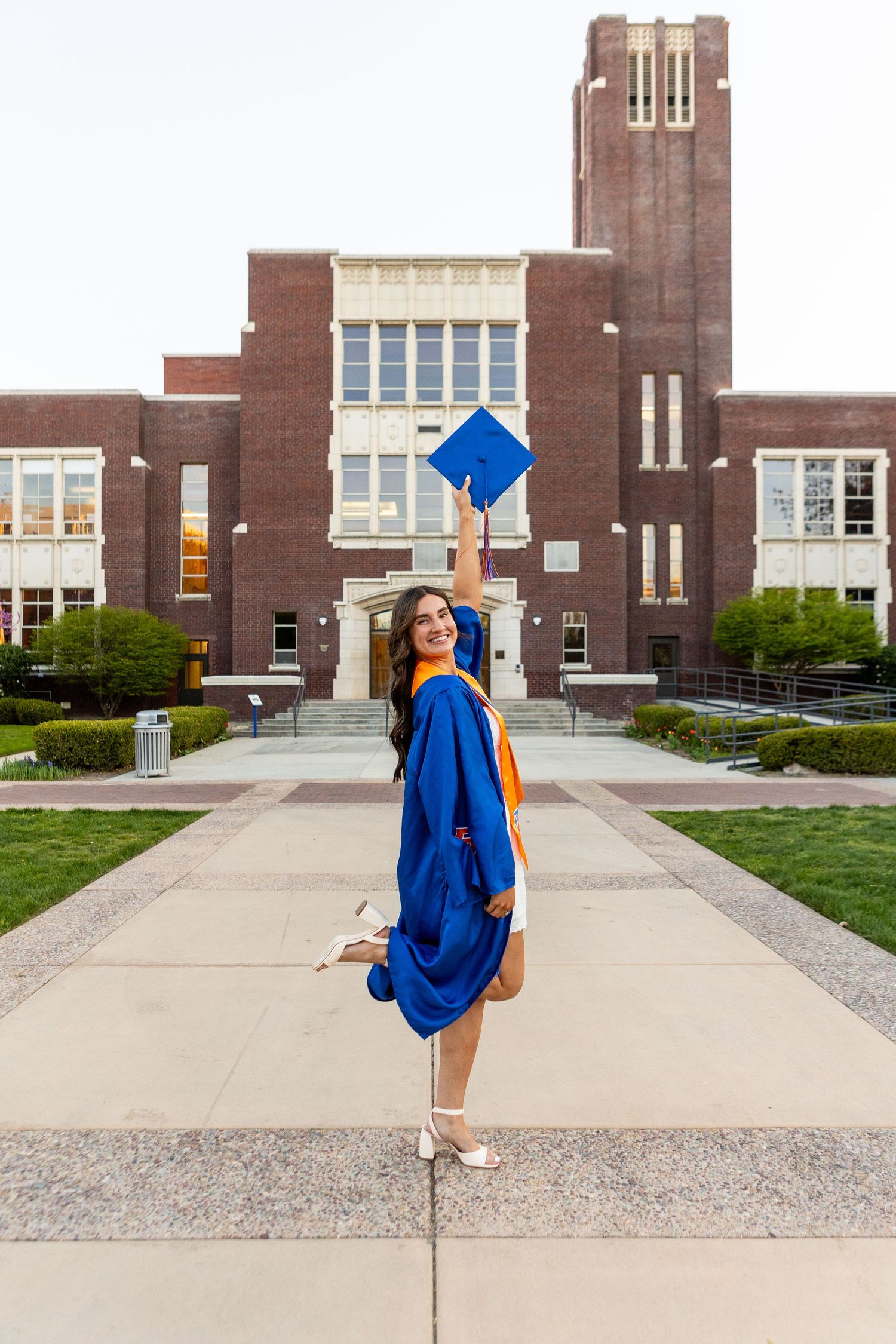 Graduate in blue and orange regalia poses in front of brick university building during photo session.