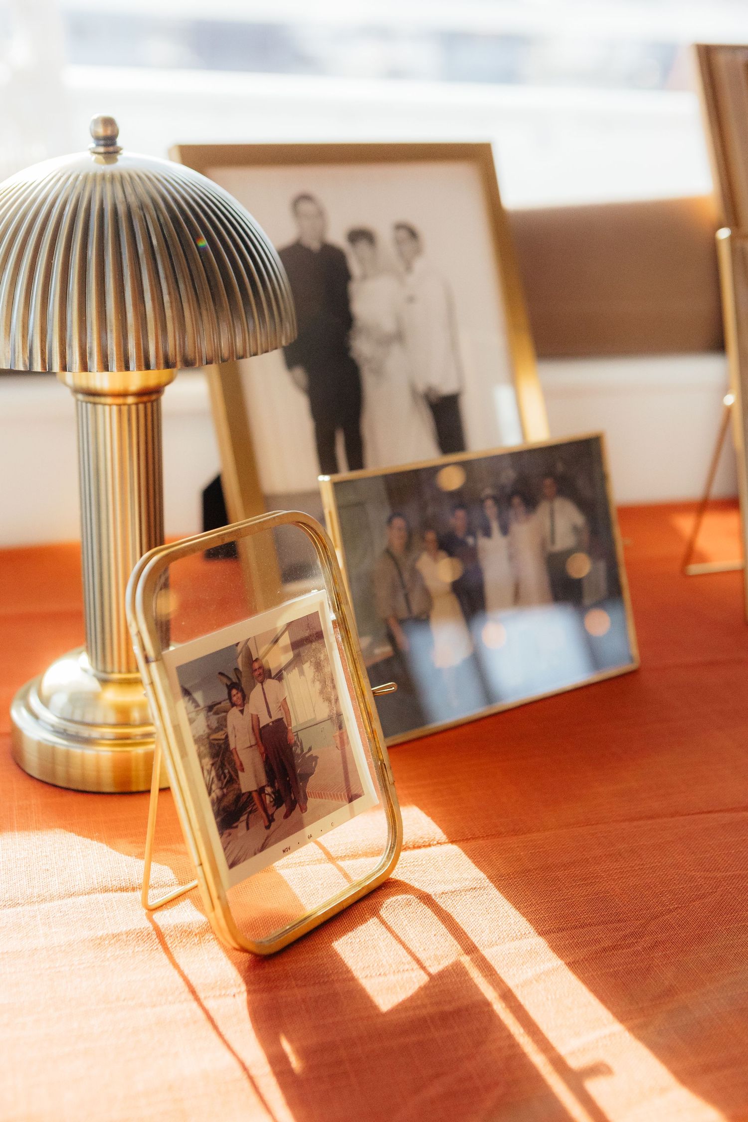 A vintage brass desk lamp illuminates framed family photographs on a wooden table in warm sunlight.