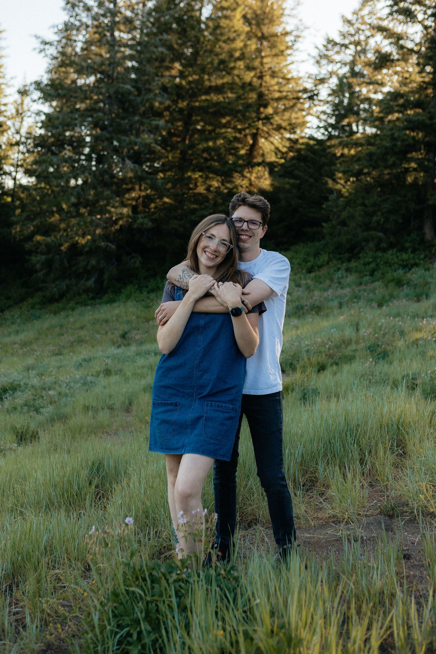 A couple embraces while standing in a grassy meadow with evergreen trees during golden hour.