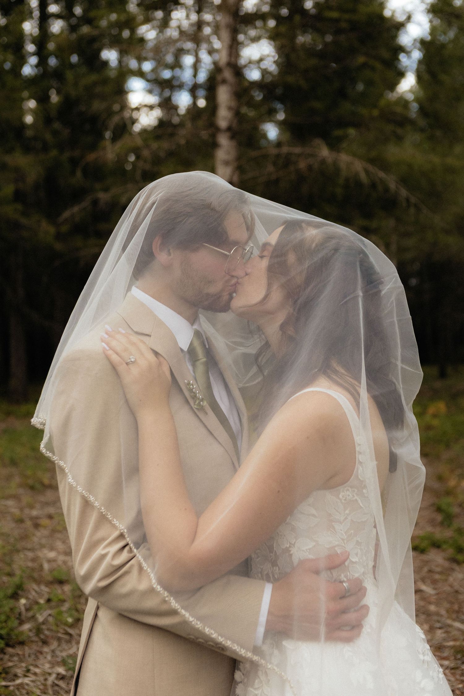 Romantic wedding couple embraces under a delicate veil in a wooded setting.