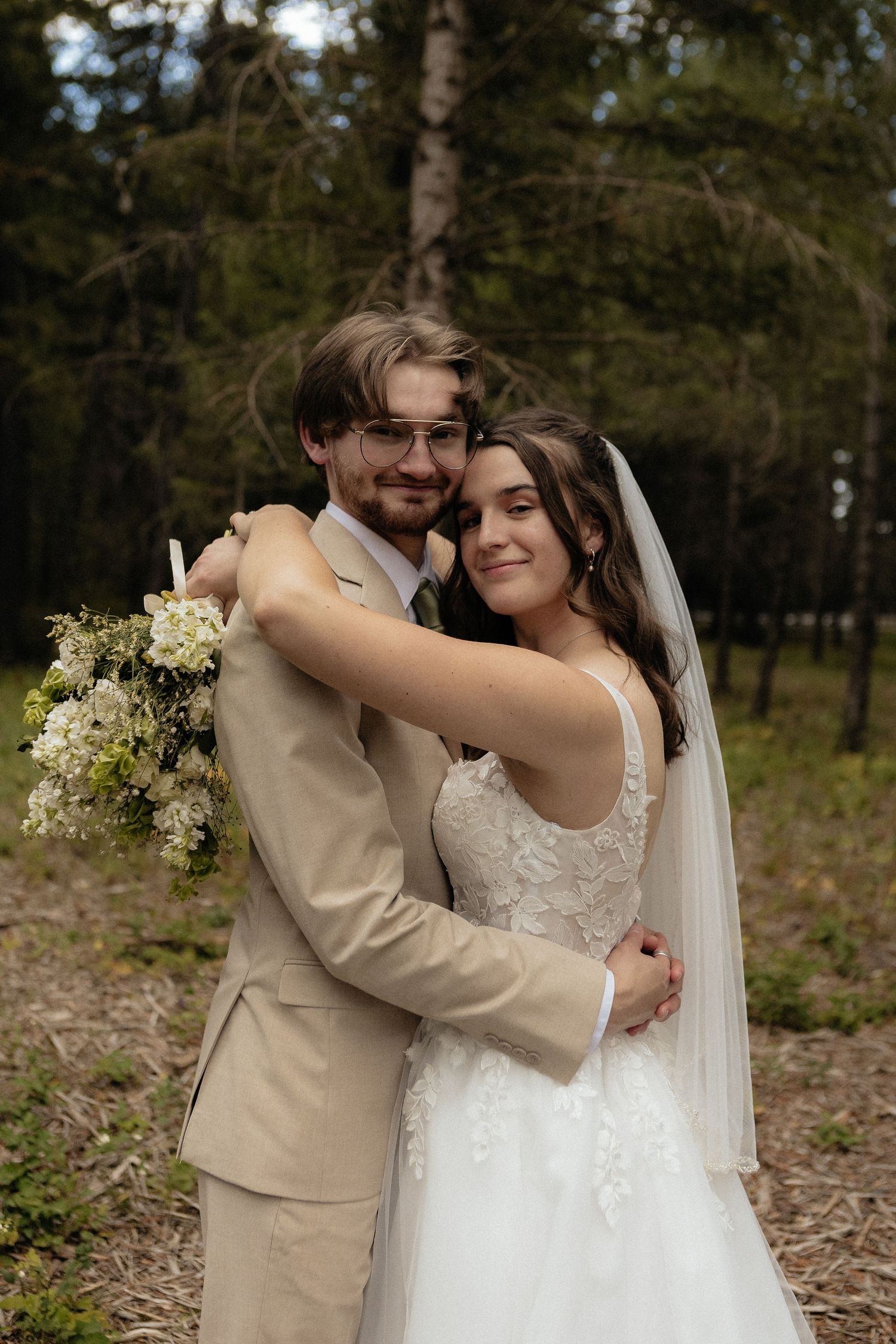 Newlyweds share an intimate moment in a forest setting with bride in lace gown and groom in beige suit.