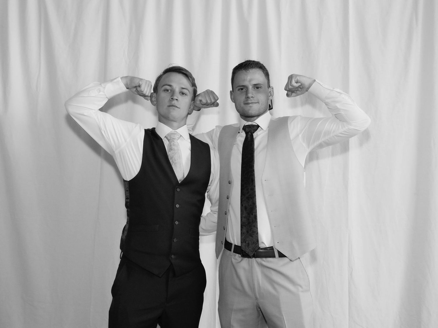 Two young men in formal attire flexing their muscles, posing for a black and white photo booth picture.