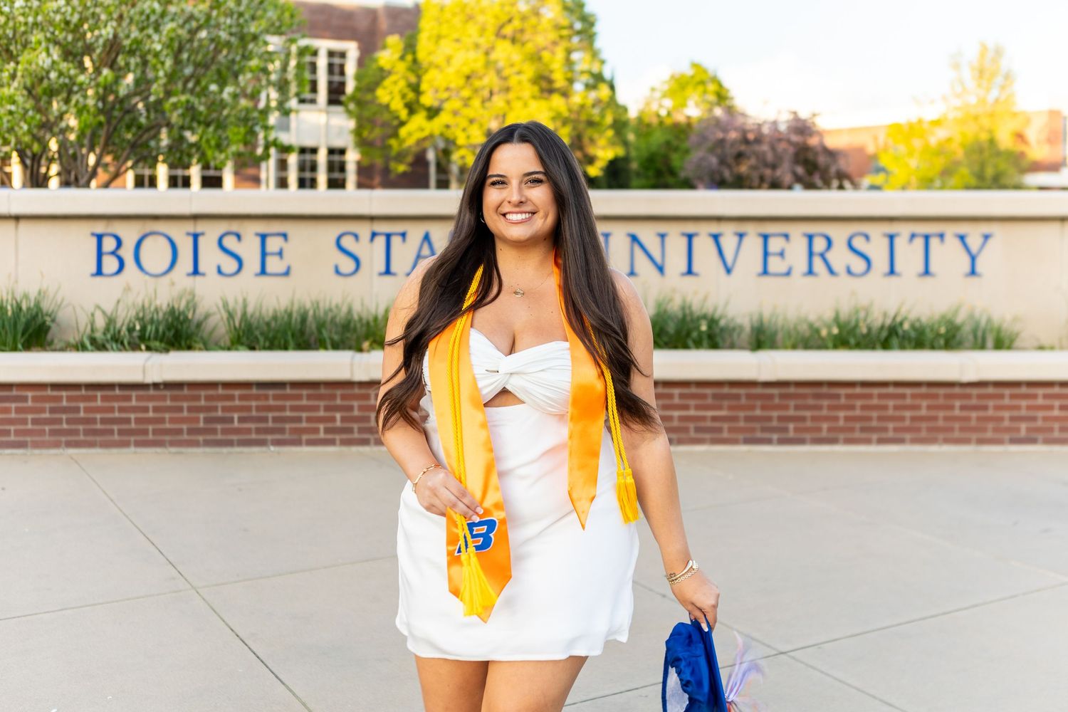 Graduate in blue and orange regalia poses on Boise State University campus during graduation photoshoot.