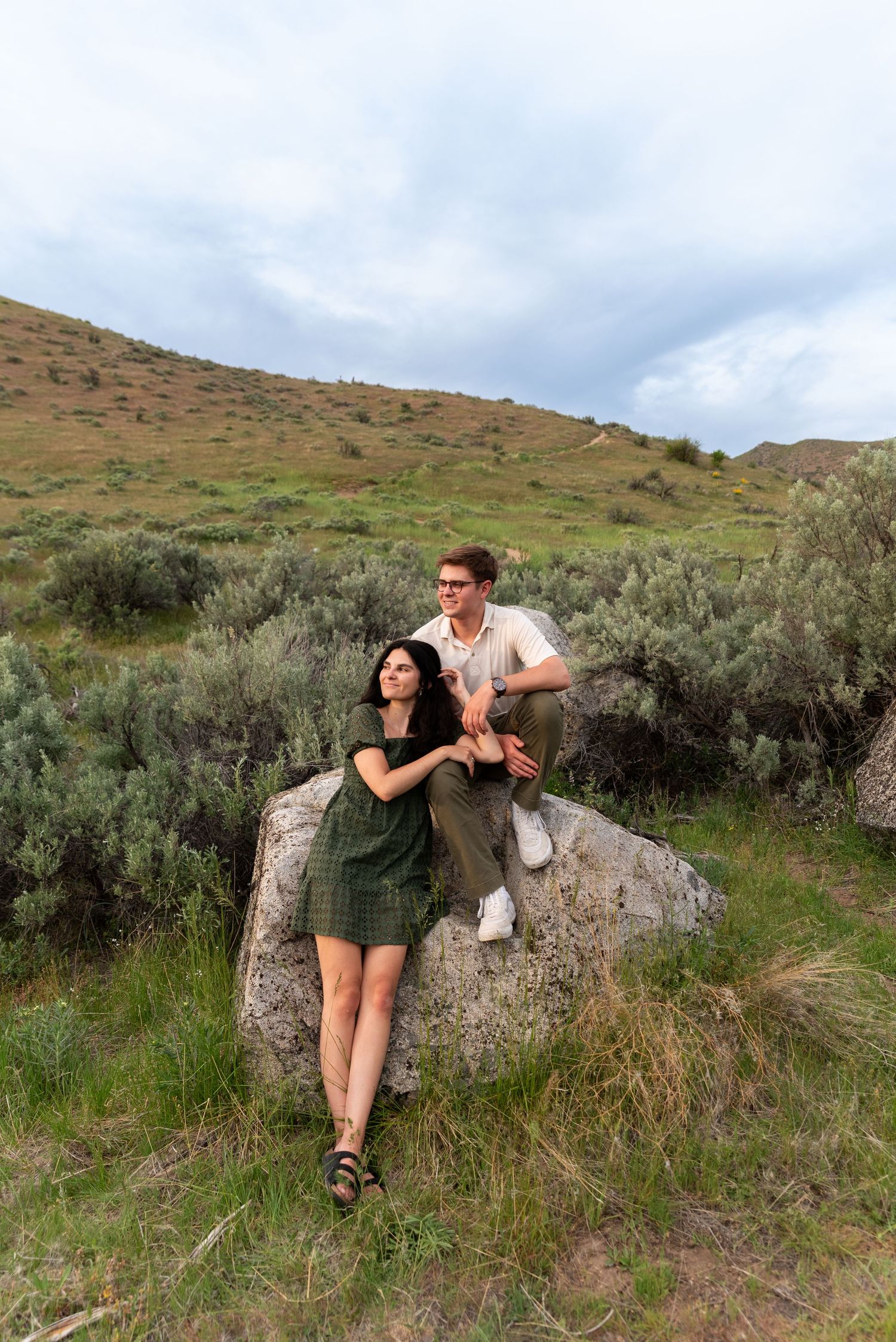 Two people posing together on a large boulder against a scenic hillside backdrop covered in sagebrush and grass.