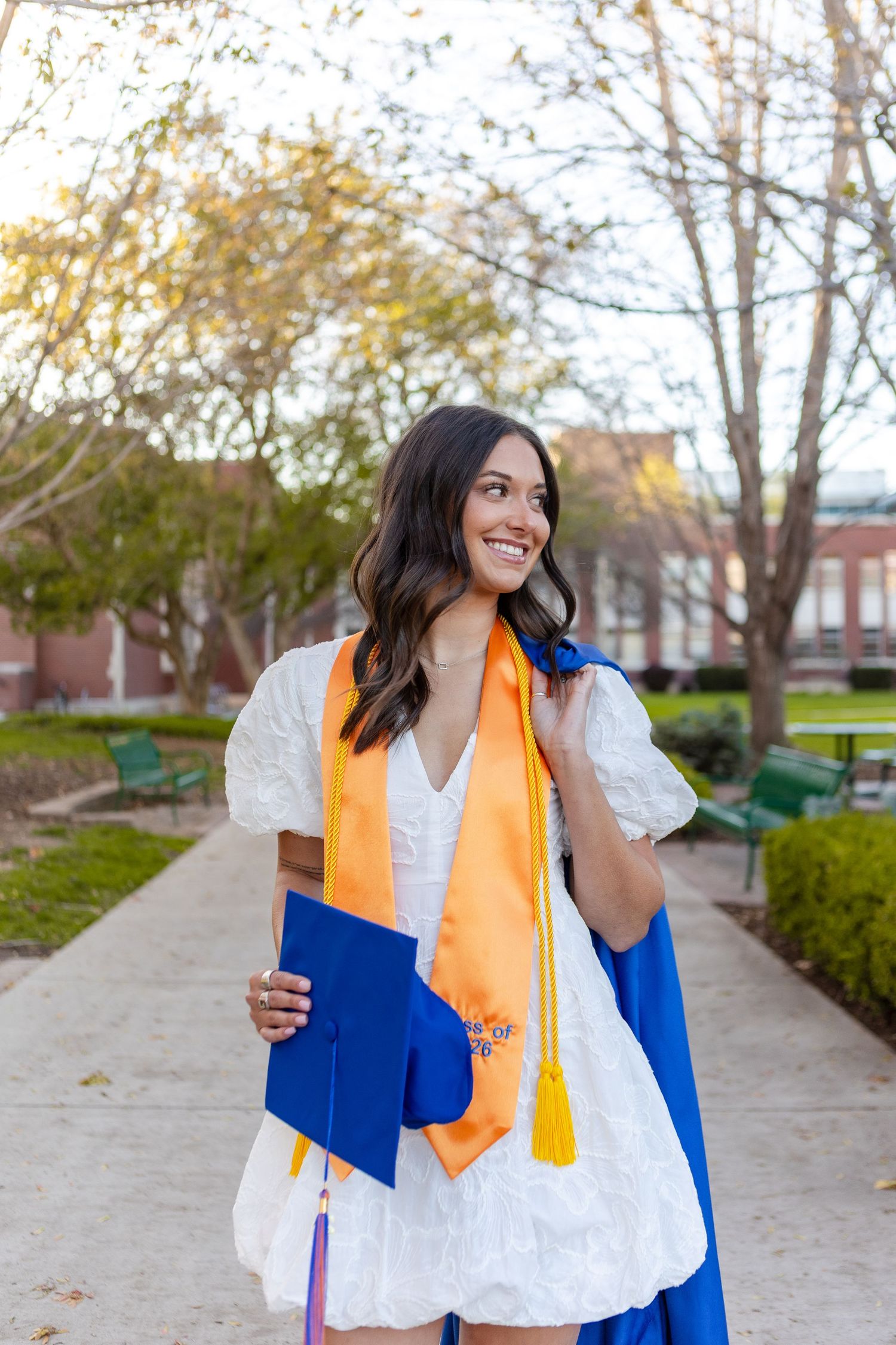 Smiling female graduate in white dress with blue cap and orange stole poses outdoors on campus pathway.