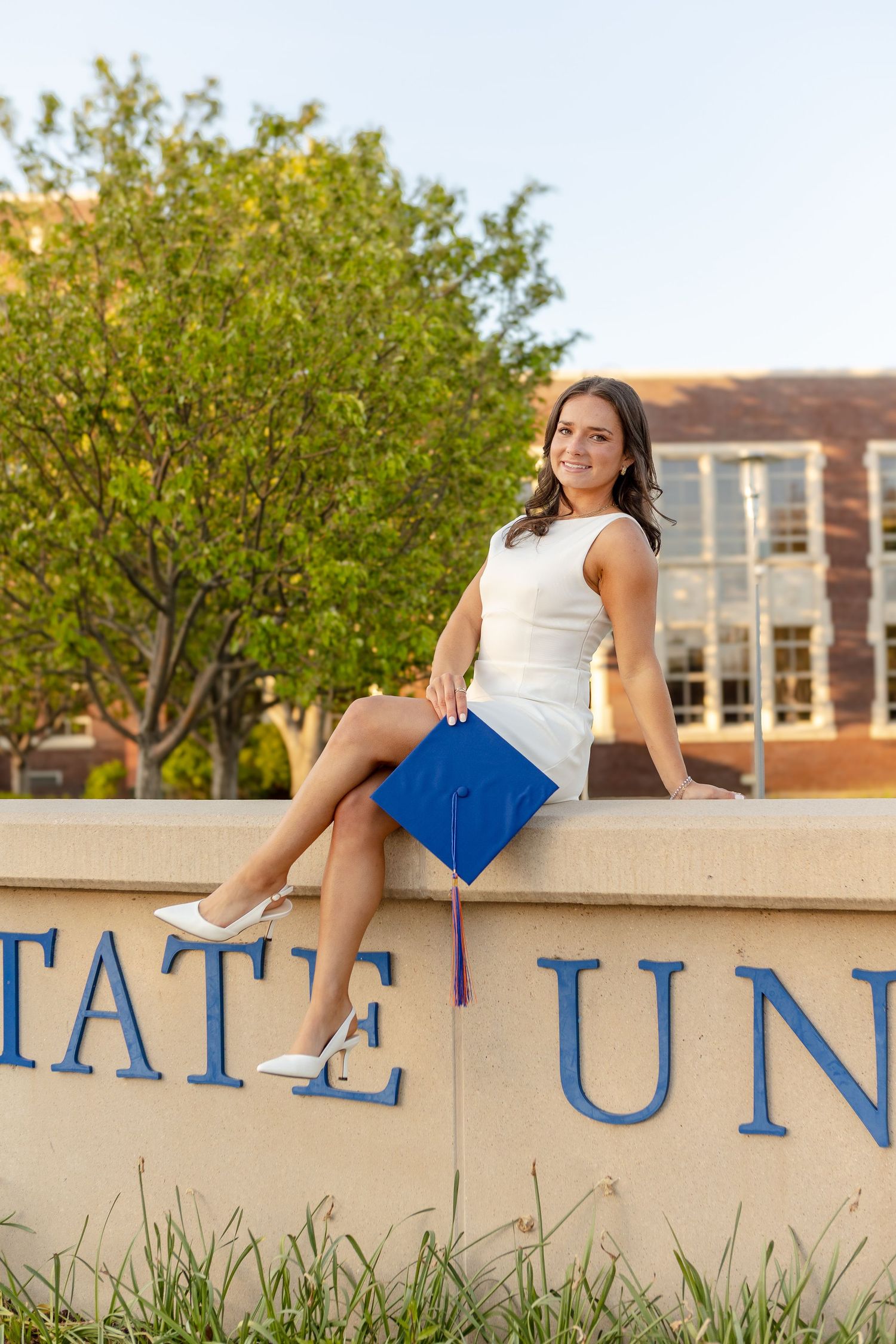 A smiling female graduate in a white dress holds a blue cap while sitting on a university sign outdoors.