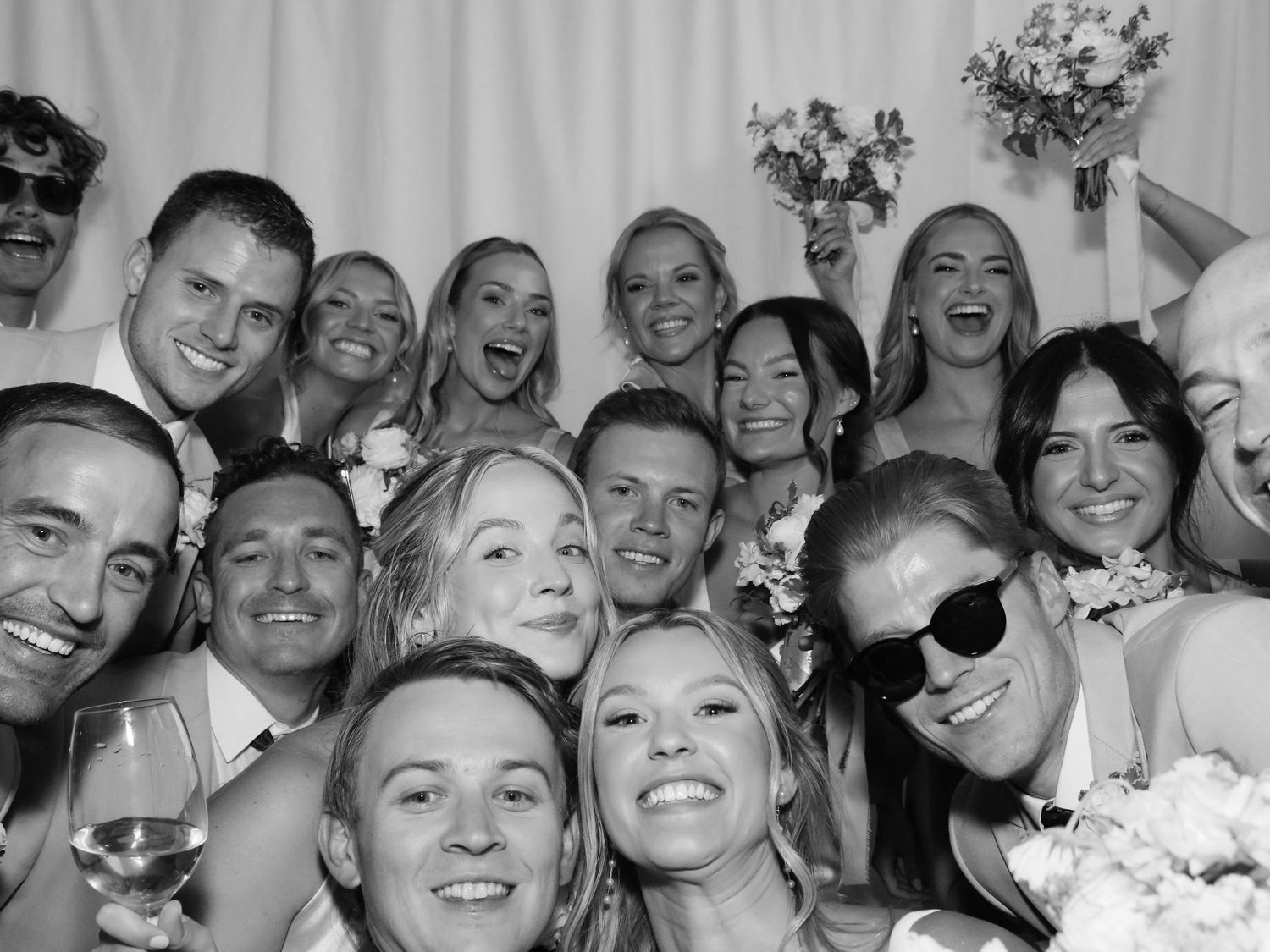 Black and white photo booth shot of a large smiling wedding party group, bride holding bouquet, celebrating together.