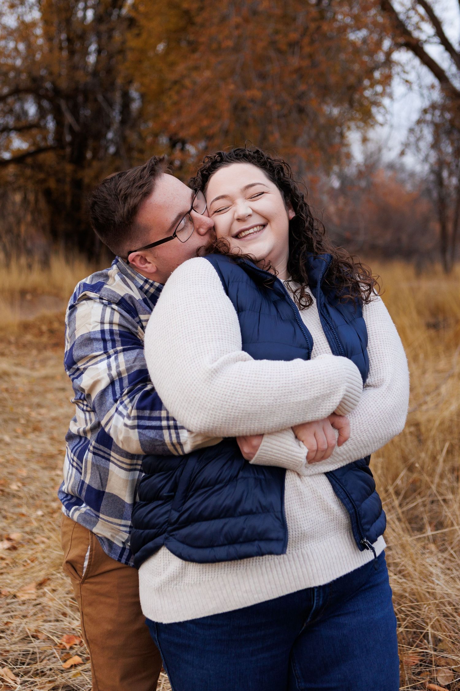 Happy couple embracing in fall field wearing coordinating cream and navy puffer vests and plaid.