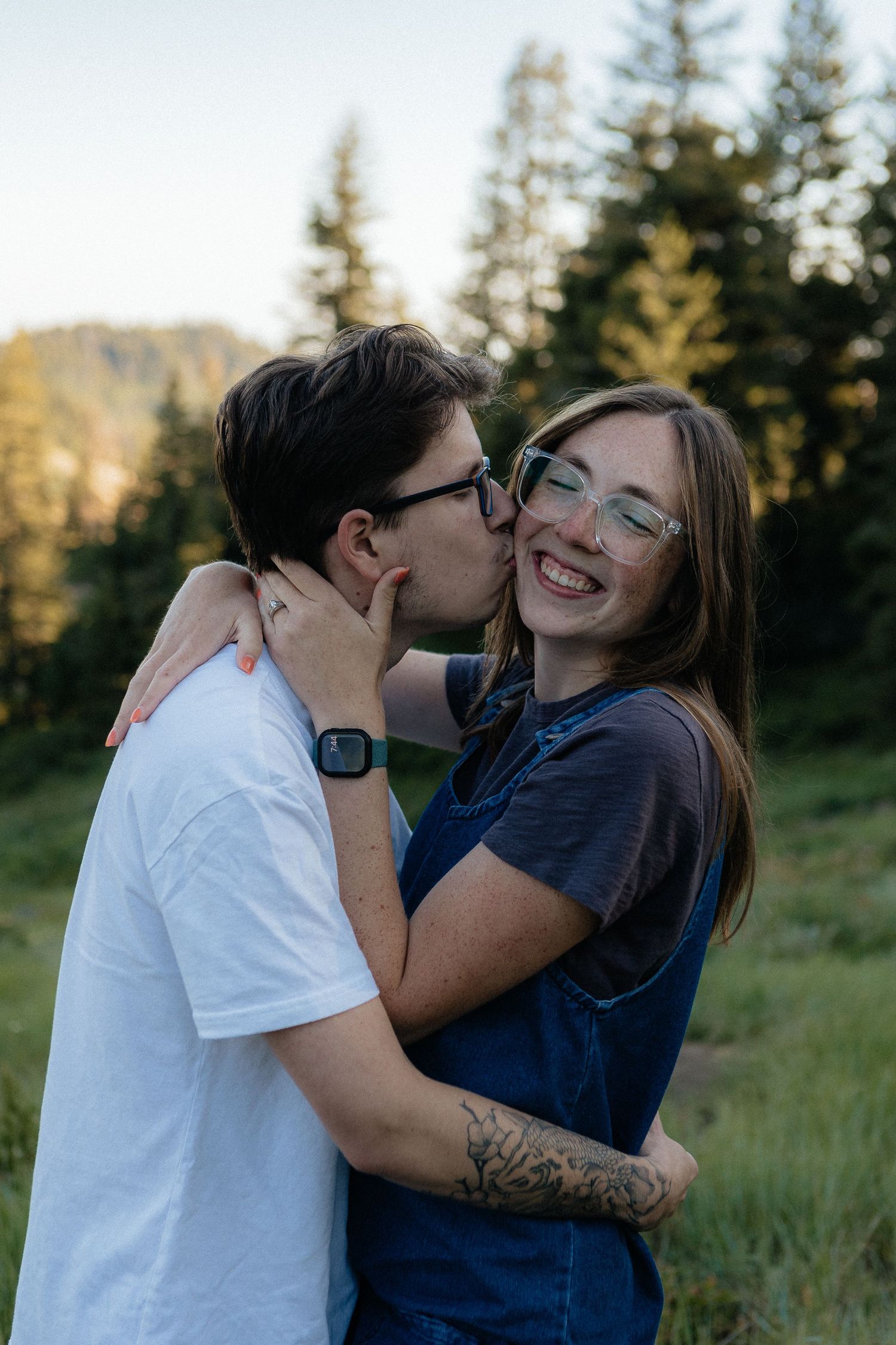 A joyful, affectionate moment captured outdoors with evergreen trees and mountains in the background.