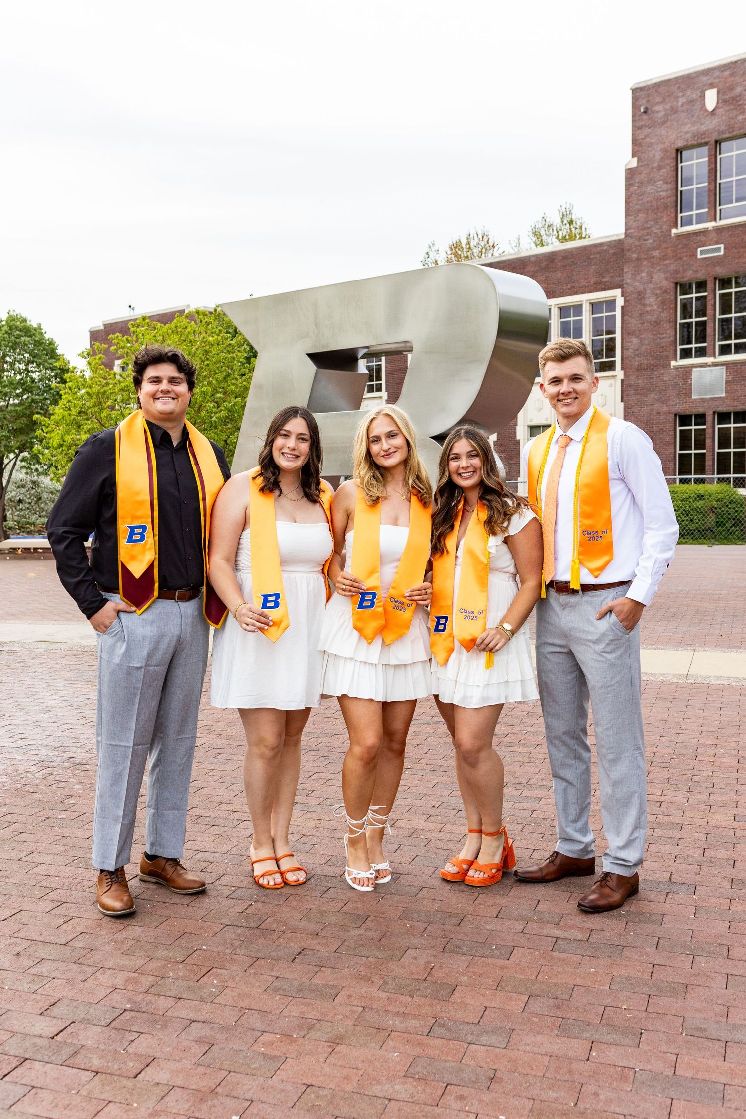 Group of graduates in orange stoles and gray suits pose together on brick courtyard.