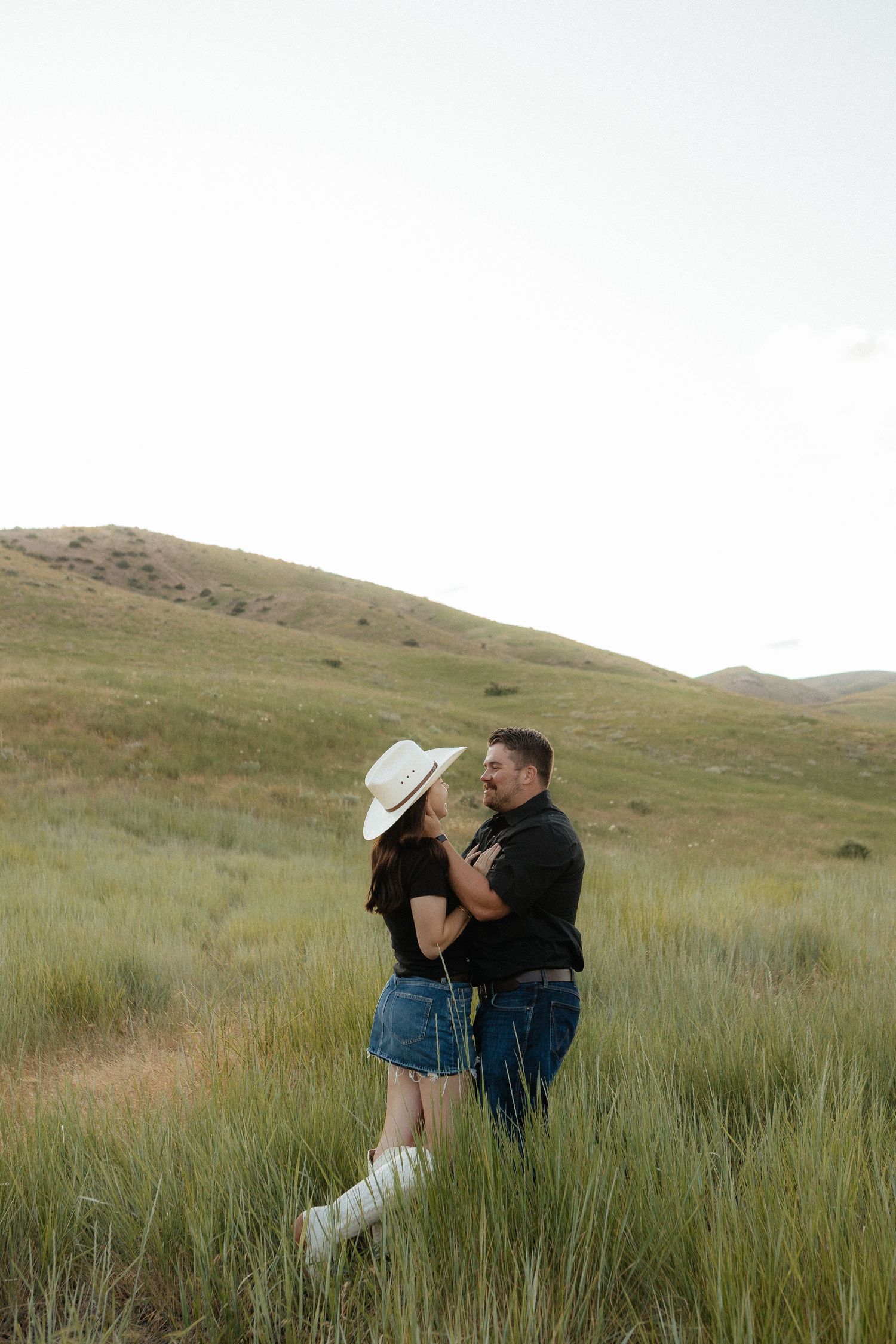 Couple embraces in a romantic photoshoot against rolling green hills and tall grass at sunset.