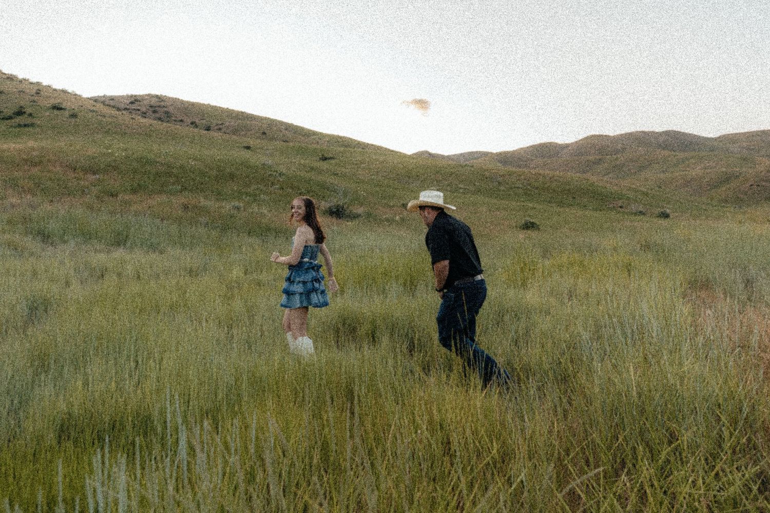 Two people walking through tall green grass in a hilly meadow during golden hour.