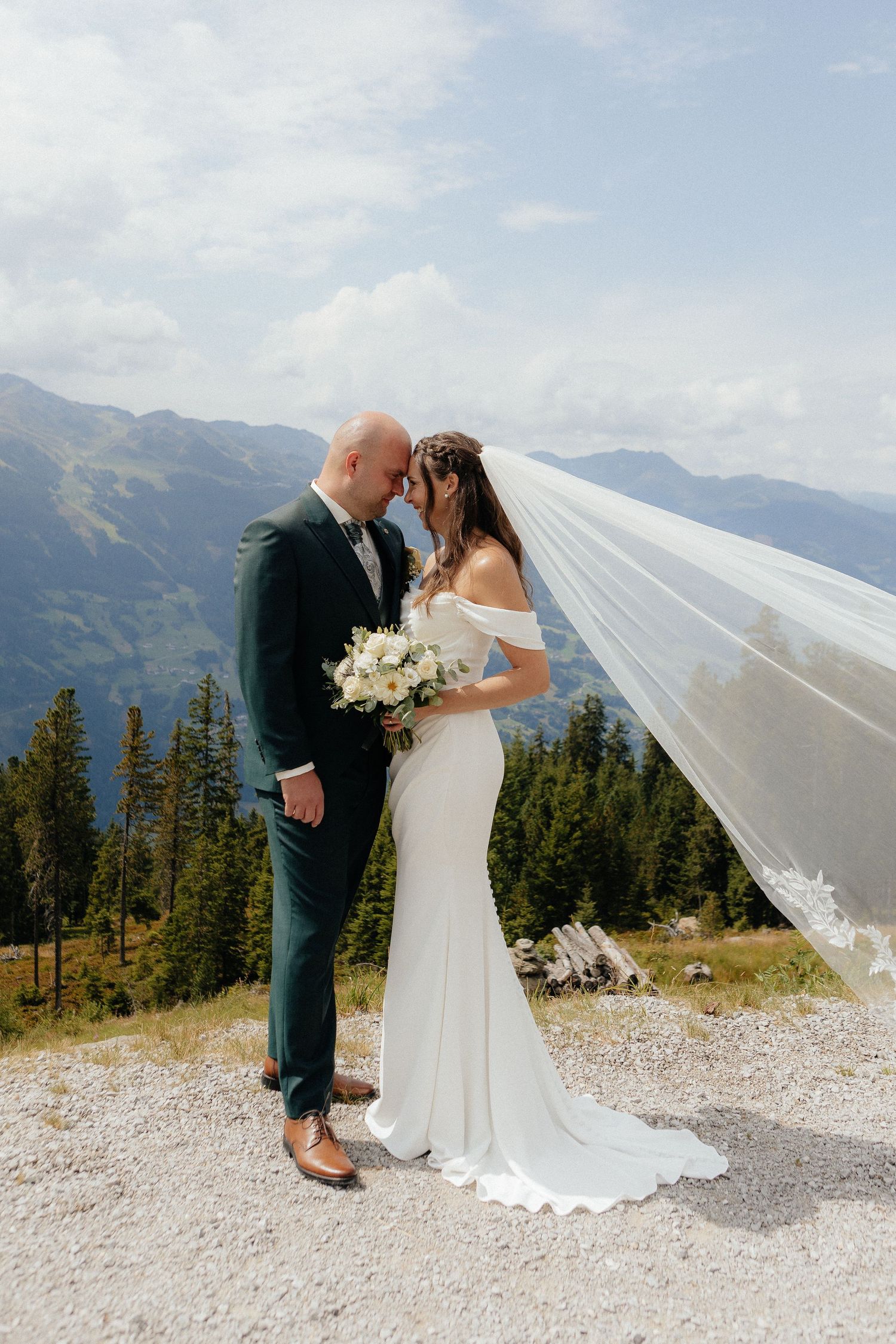 A bride and groom share a romantic moment on a mountaintop with a flowing veil catching the breeze.