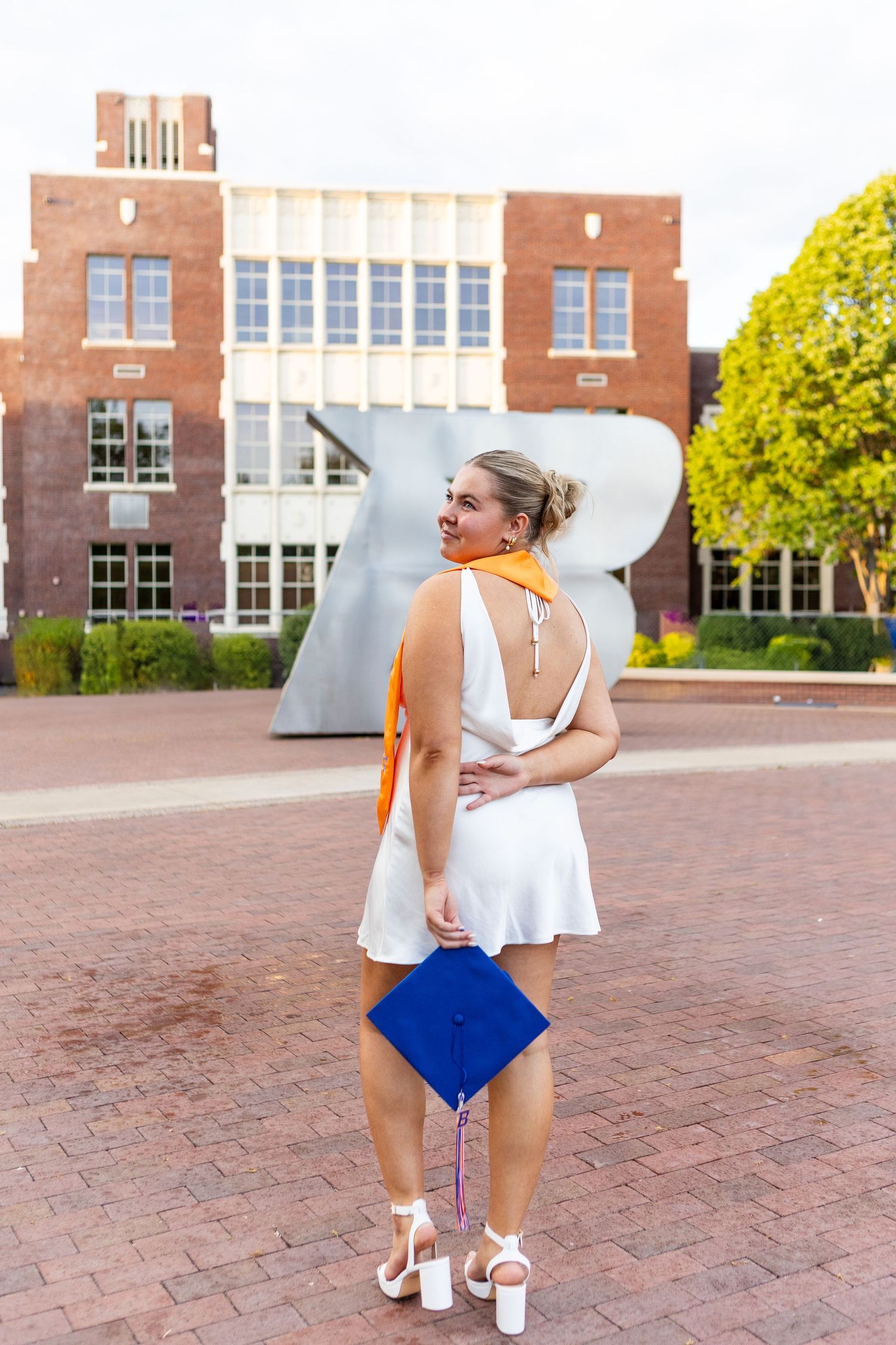 Graduate in white dress poses near brick building with blue graduation cap.