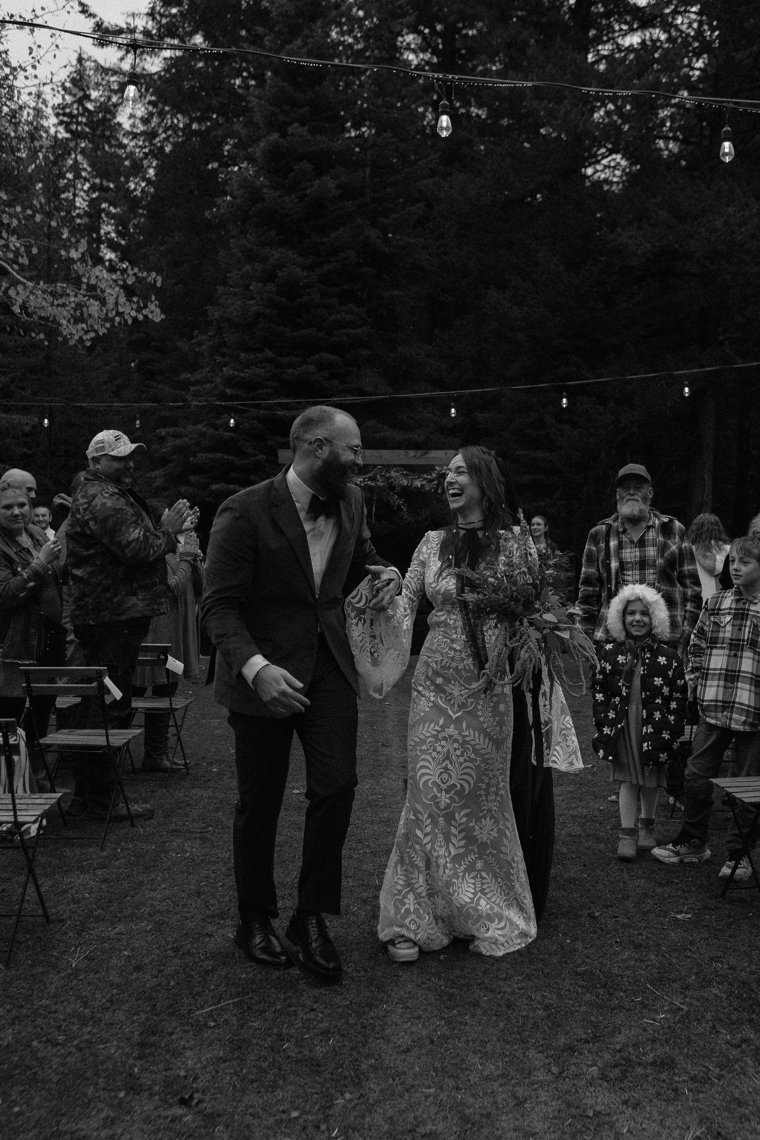 Black and white capture of wedding reception guests gathering under string lights at dusk.