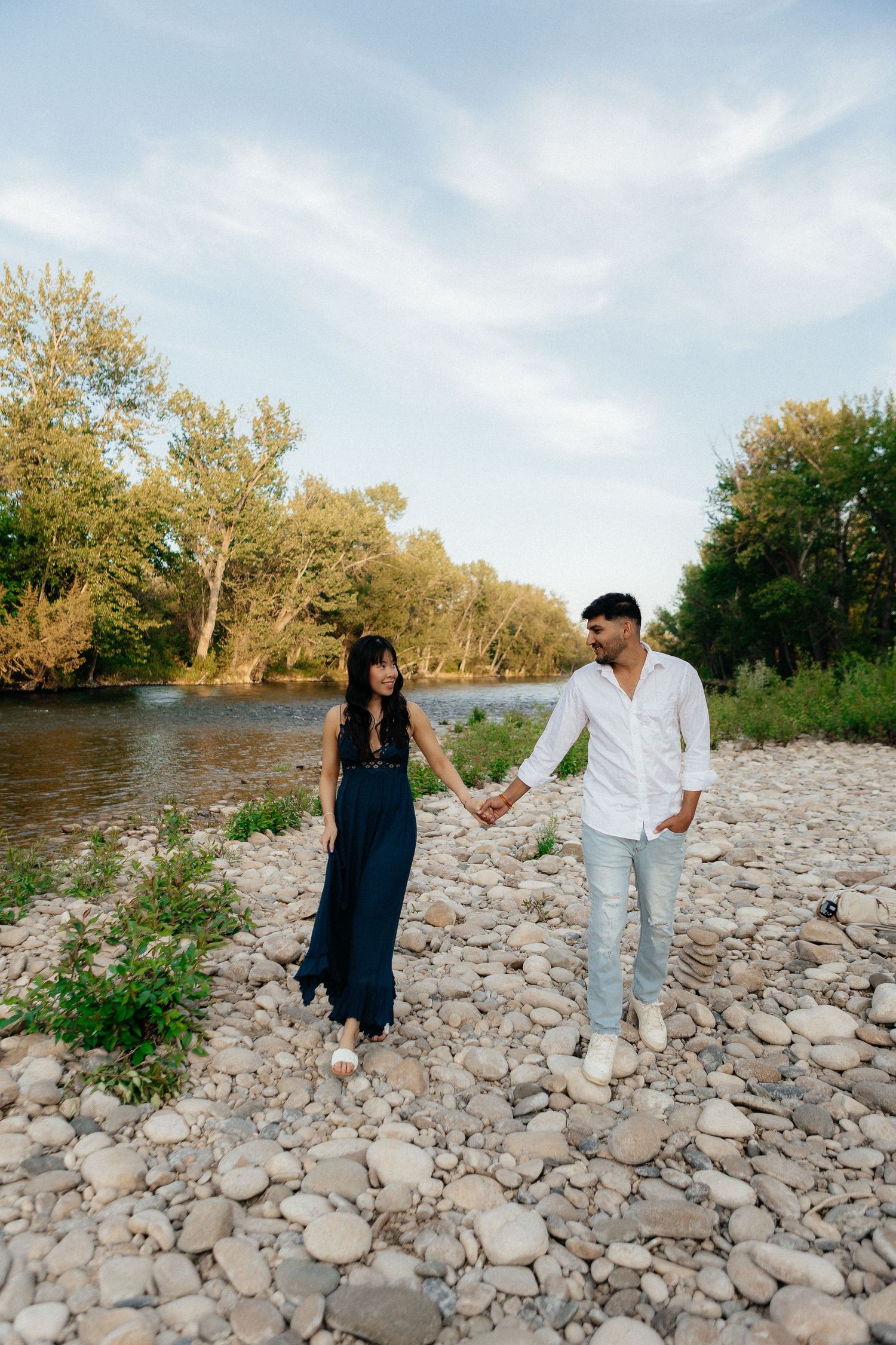 A couple walks together on a rocky riverbank surrounded by autumn trees during a sunny day at sunset.