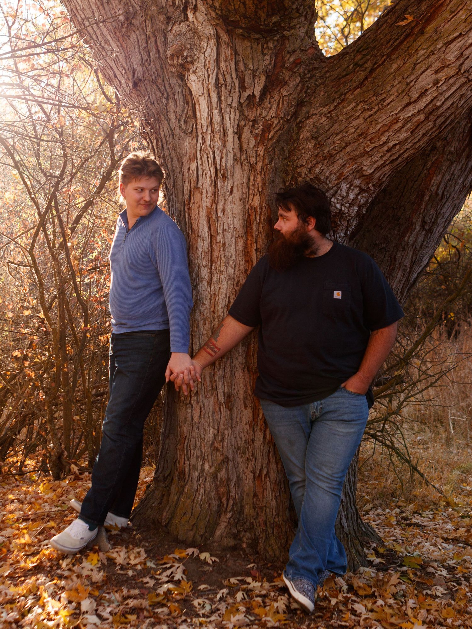 Two people stand beside a large tree trunk with autumn foliage scattered on the ground during fall season.