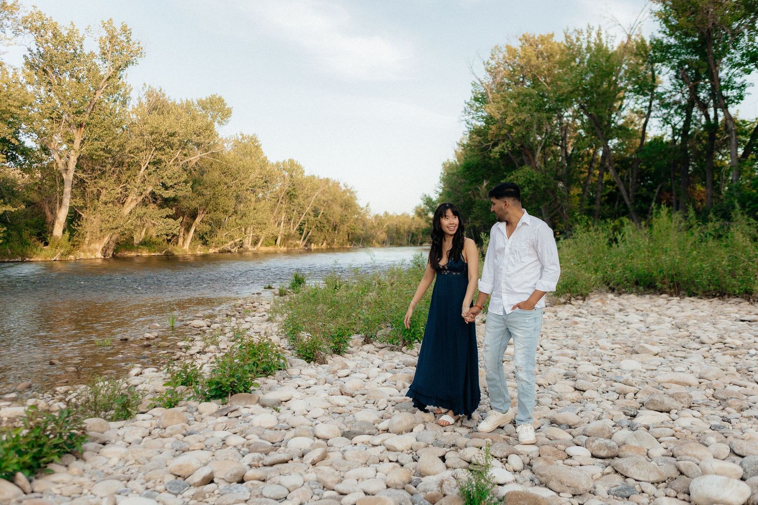 A romantic couple walks along a scenic rocky beach shoreline surrounded by trees during a summer evening.