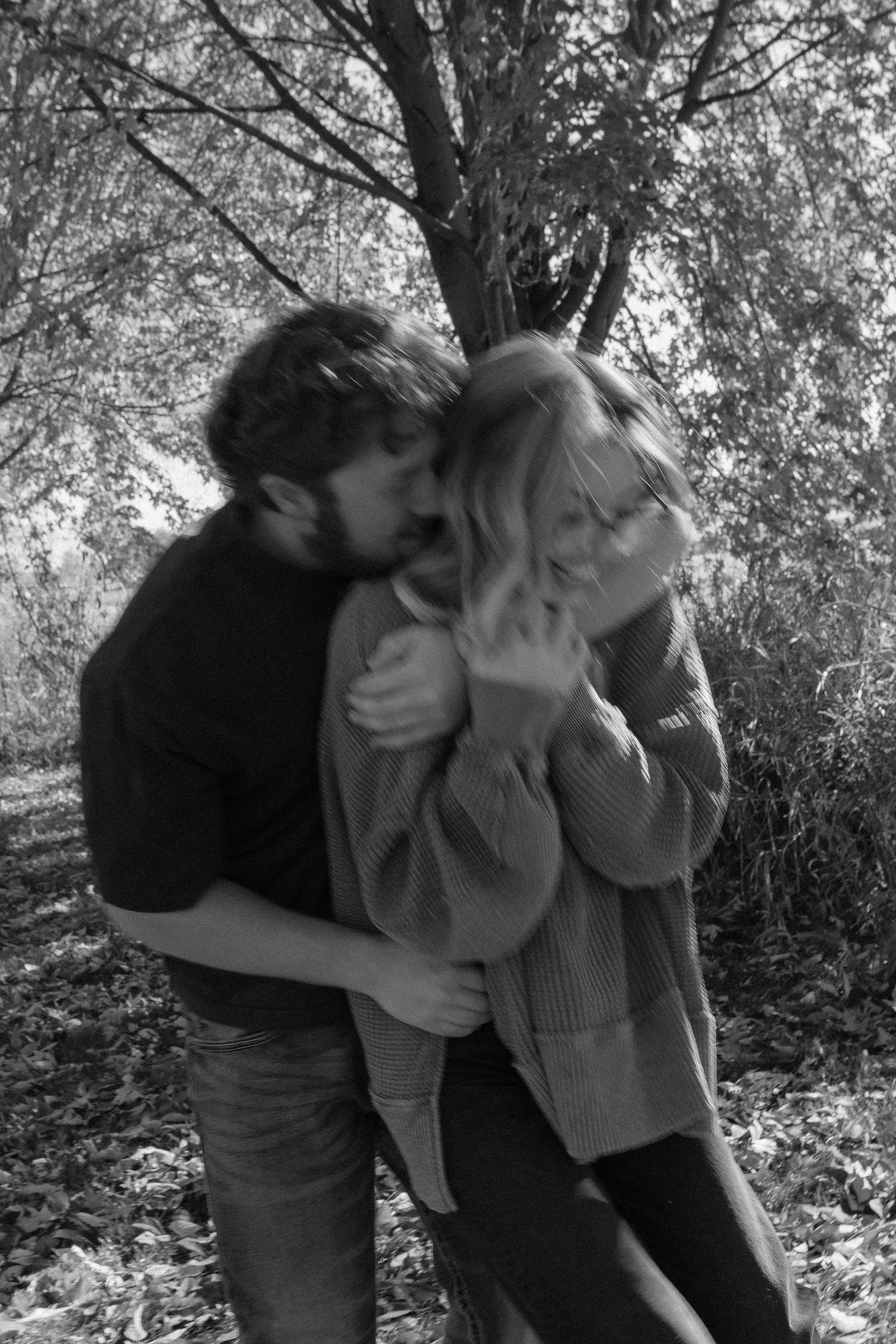 A romantic couple shares an intimate moment under trees in black and white photography.