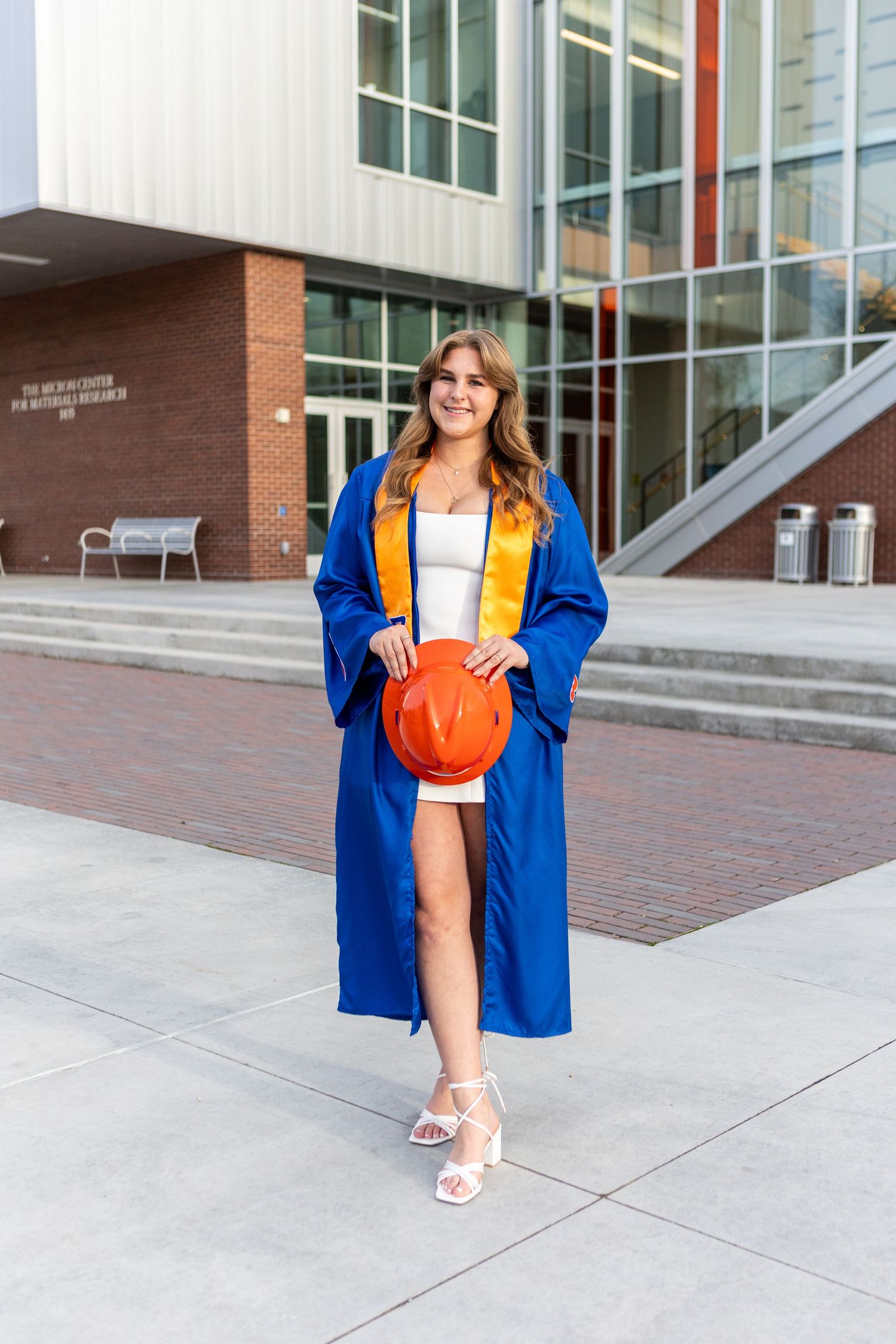 Graduate in blue cap and gown poses playfully outside modern university building.