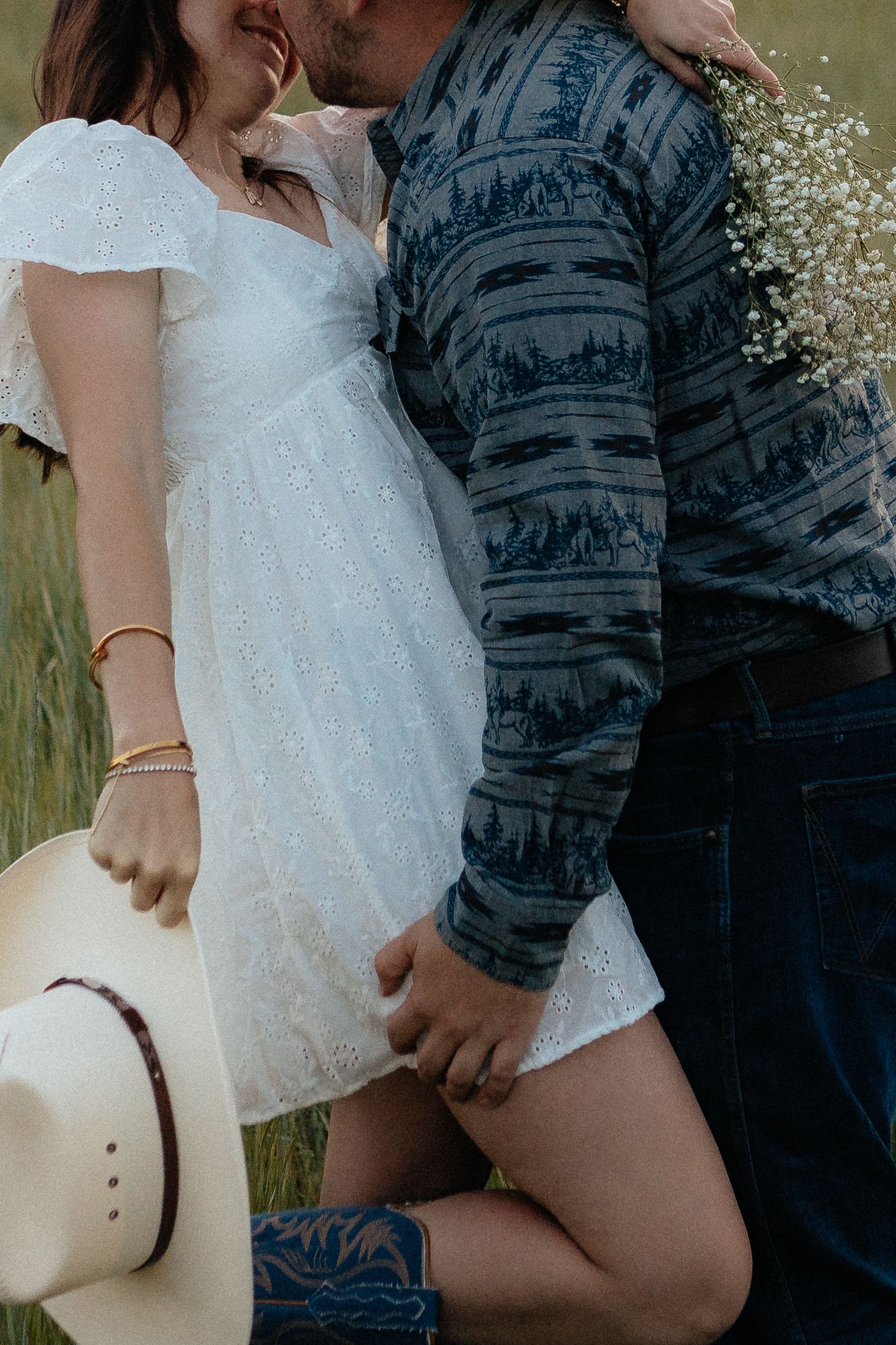 Romantic couple embracing in a field, with white dress, blue striped shirt and cowboy hat details in summer setting.