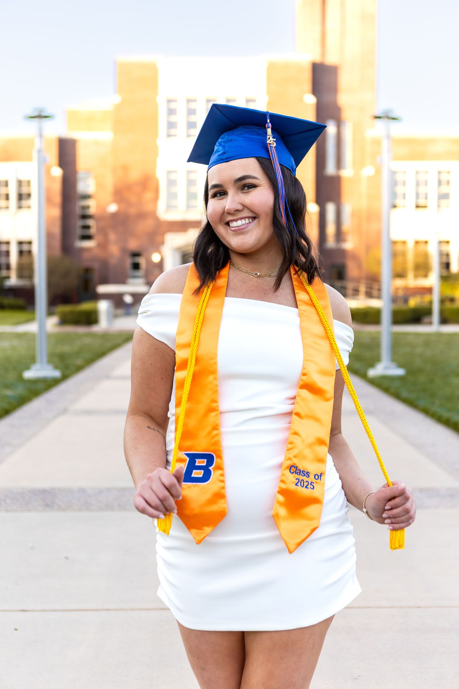 Graduate wearing white dress and orange stole poses with diploma on campus grounds.